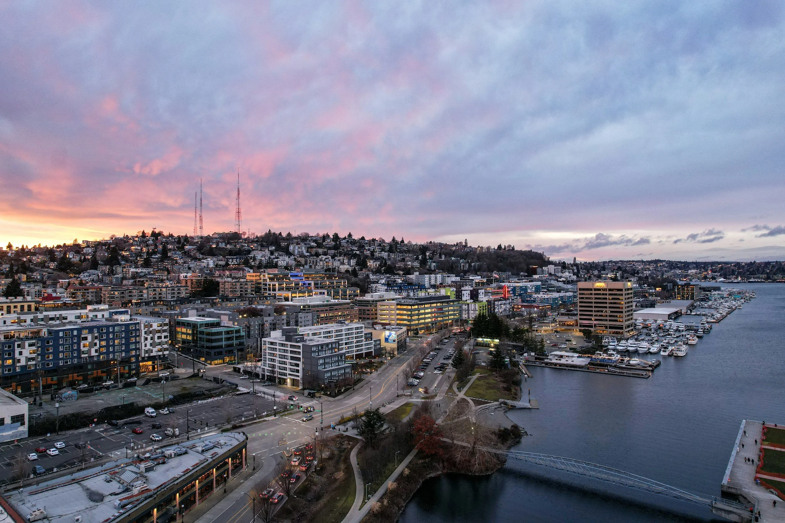 Cityscape at dusk with colorful sunset sky, modern buildings, a marina filled with boats, and a waterfront park.