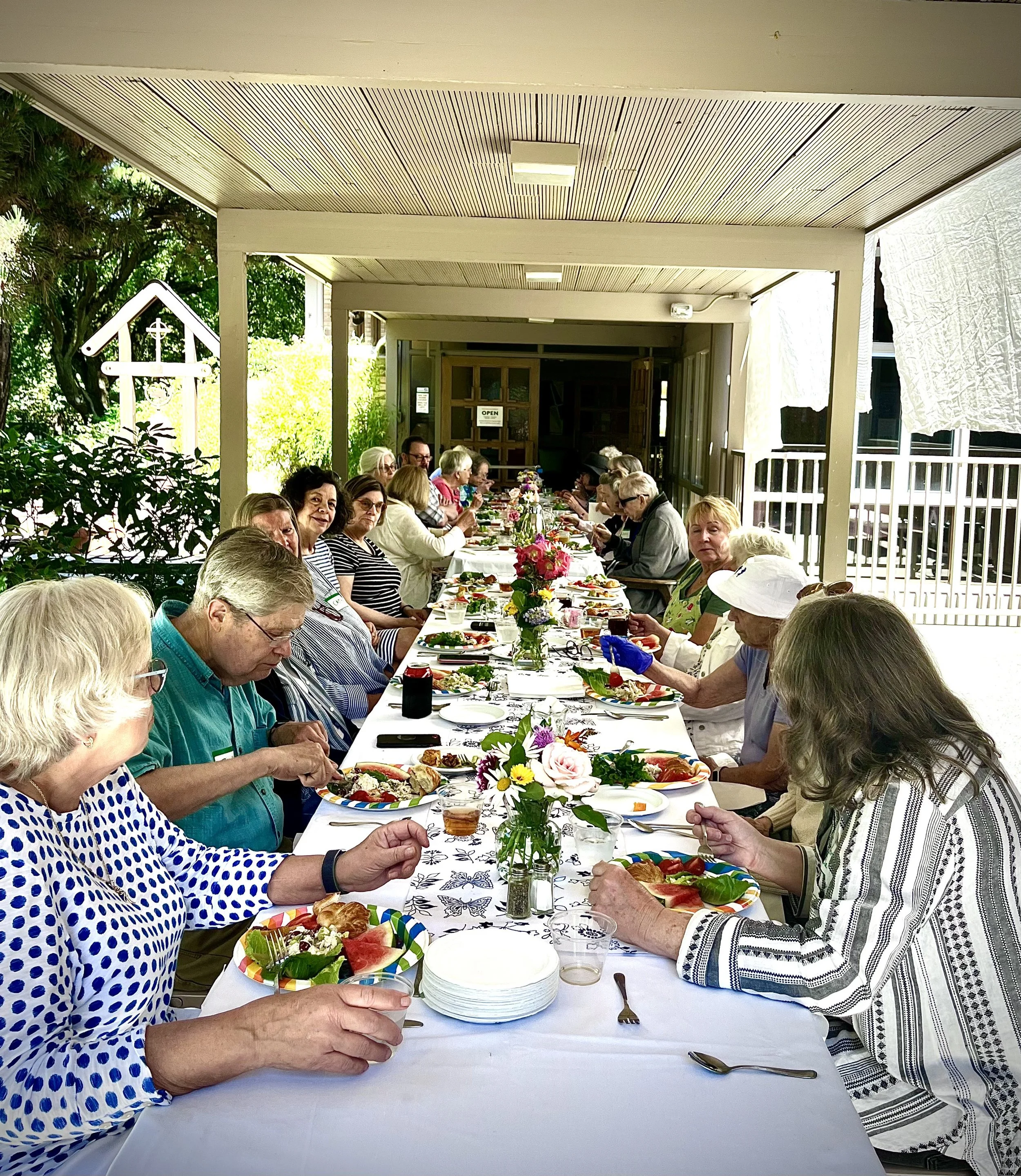 A large group of elderly people sitting at a long outdoor table decorated with flowers, enjoying a meal together under a covered patio with greenery in the background.