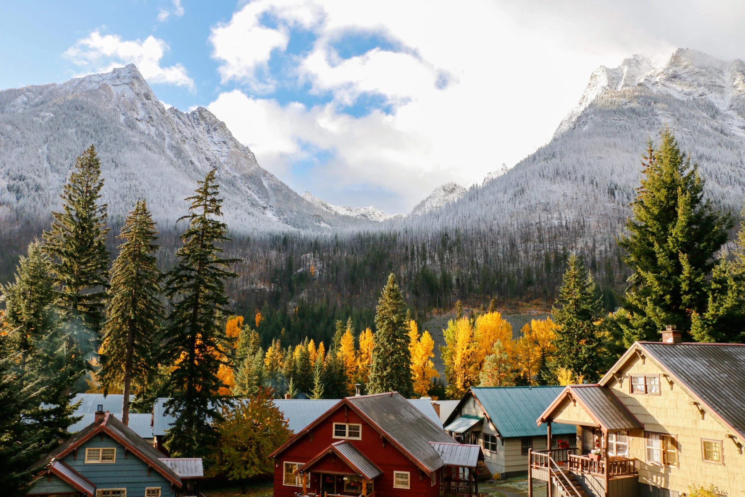 Mountain valley scene with snow-capped peaks, tall evergreen trees with yellow foliage, and colorful houses in the foreground.