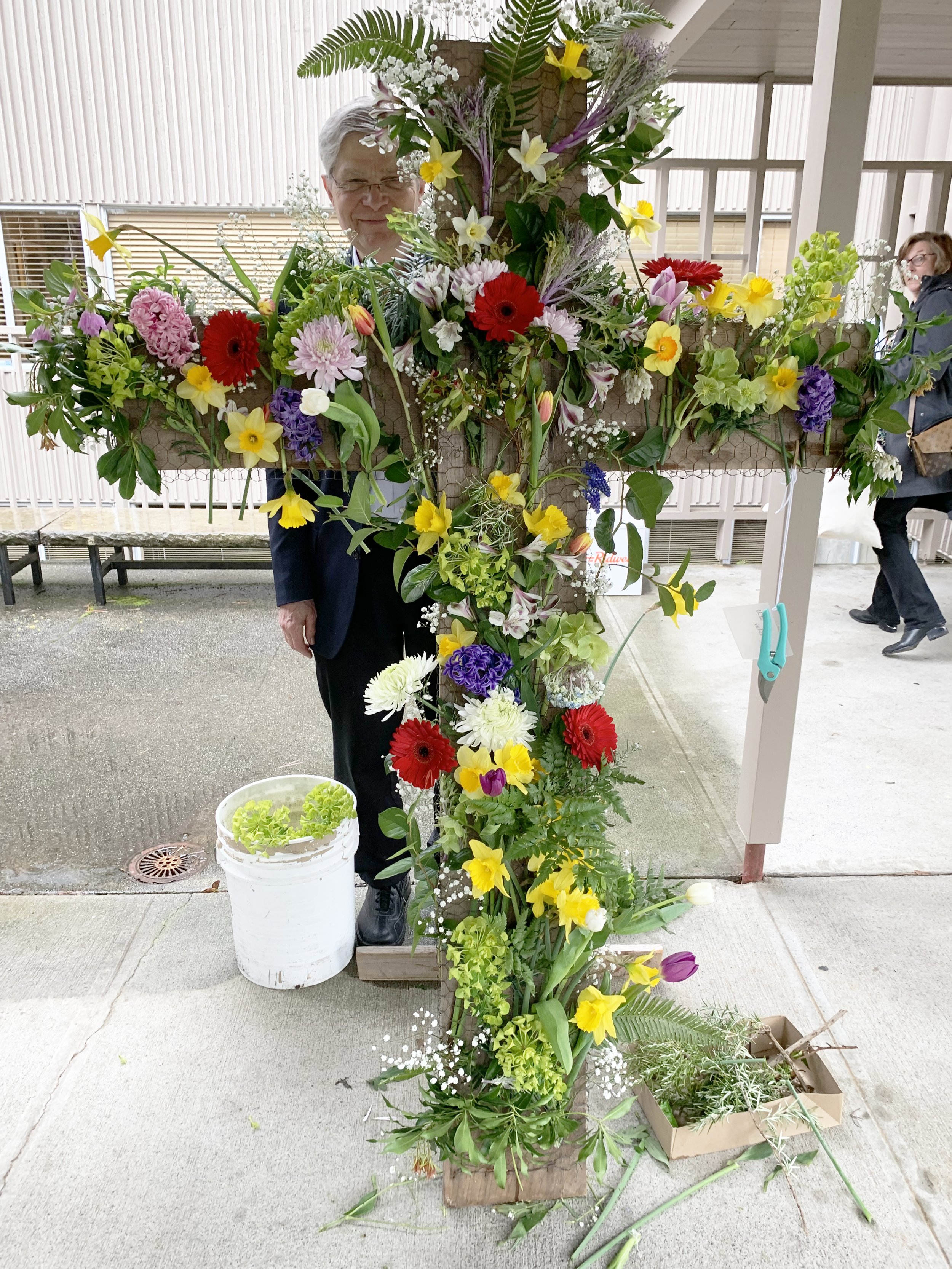 A man standing behind a large cross-shaped floral arrangement made of colorful flowers including daffodils and tulips, with another person partially visible in the background.