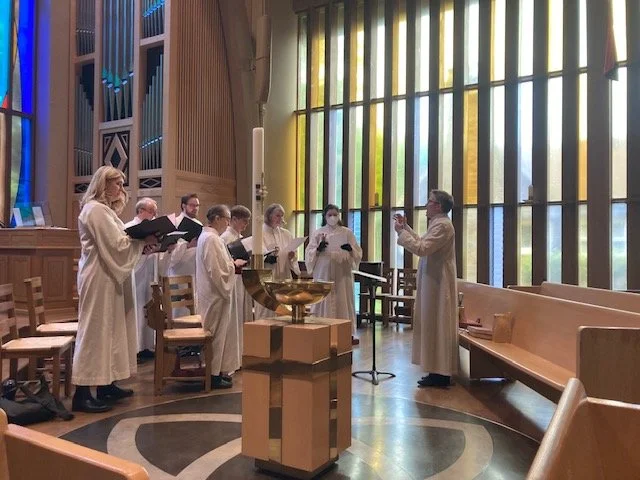 A choir of children and a woman singing in a church with tall stained glass windows, wooden pews, and an organ in the background.