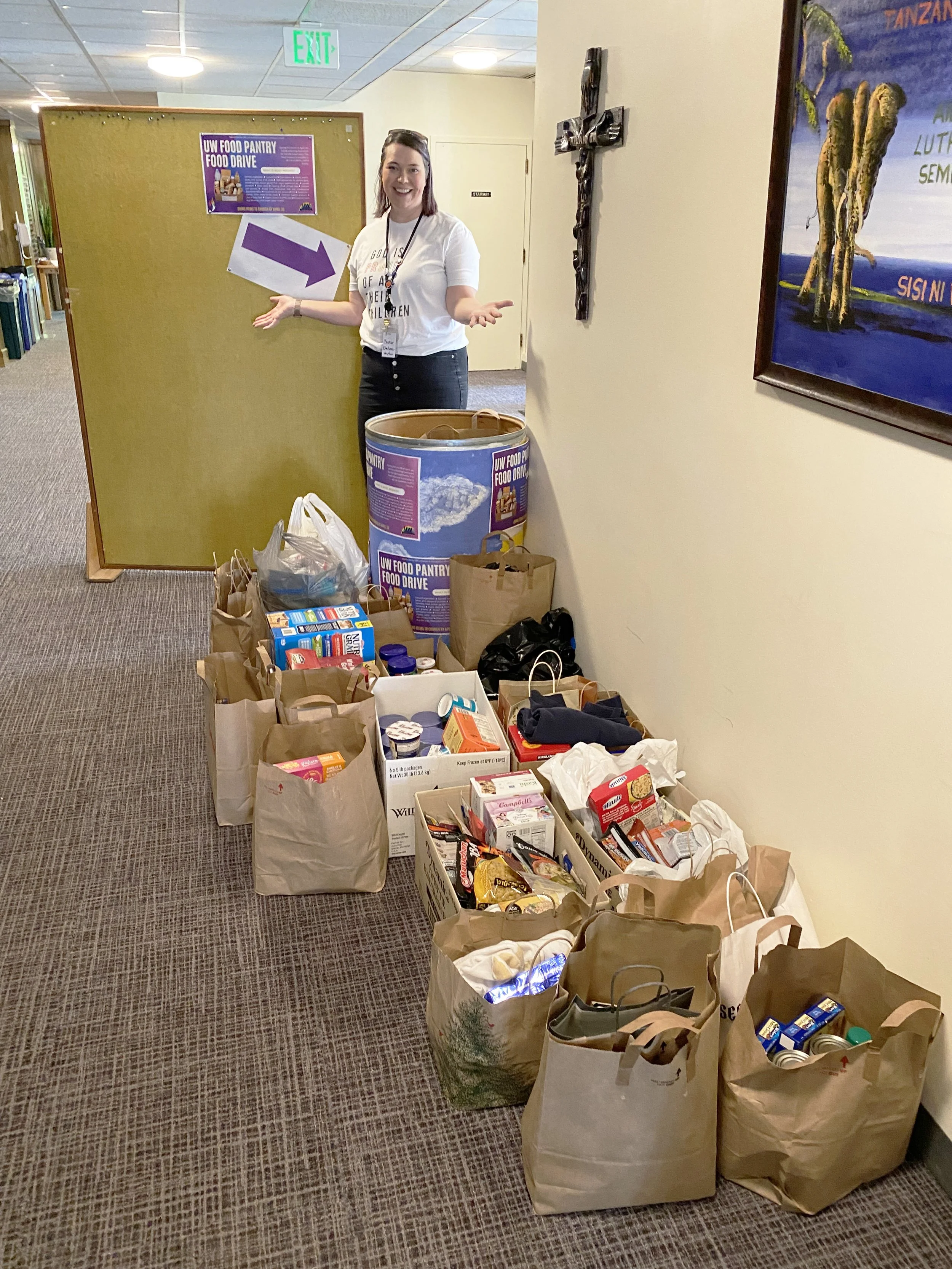 A woman standing next to a table of grocery bags filled with food in a hallway with wall art and a bulletin board.