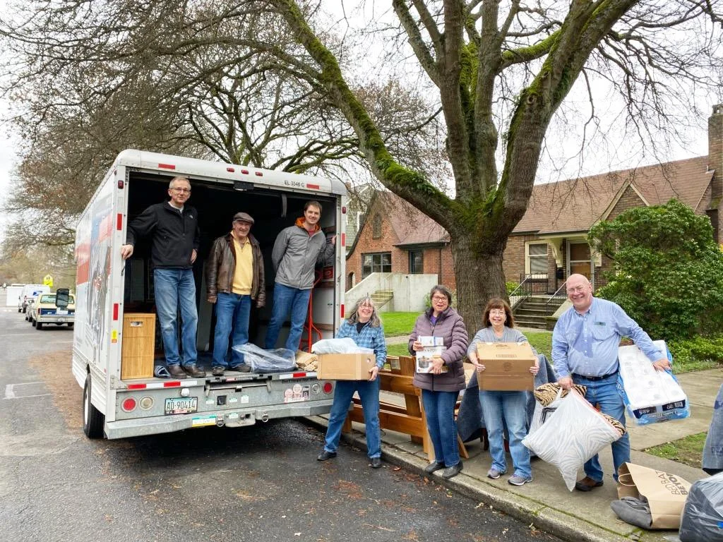 A group of people unloading items from a moving truck onto a sidewalk. They are holding boxes, bags, and household items. The truck is parked on a residential street with trees and houses in the background.