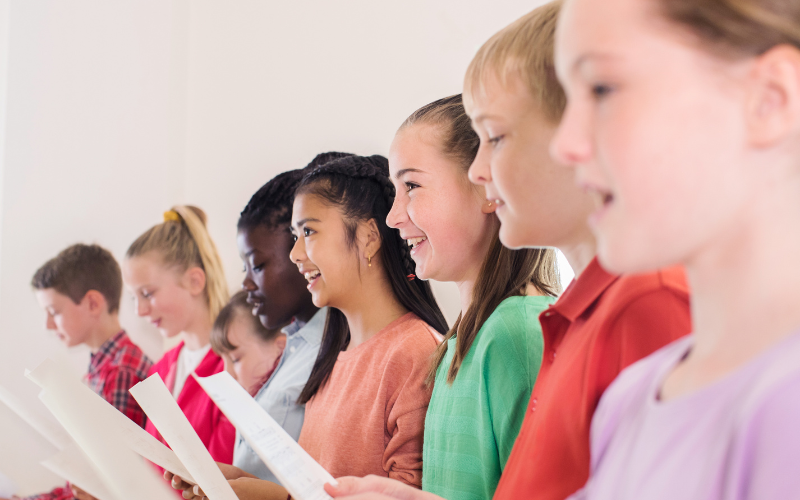 A group of children standing in a line, holding papers, smiling, and looking to the right.