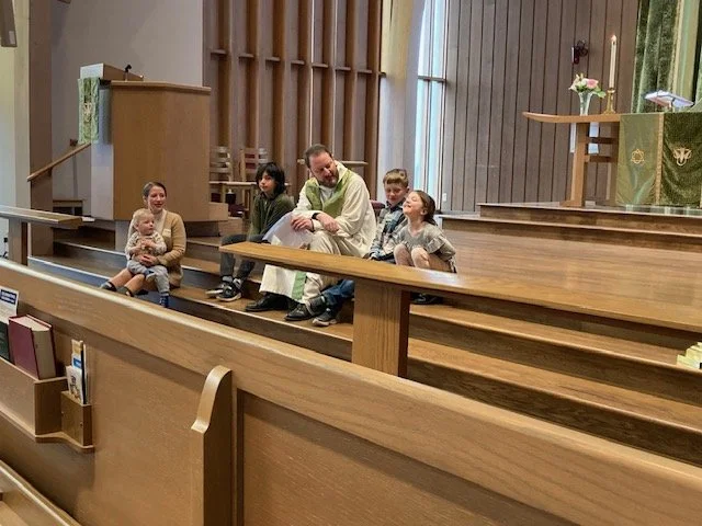 A priest or minister sitting on a wooden step in a church with five children, some sitting and some standing around him, near the altar with flowers.