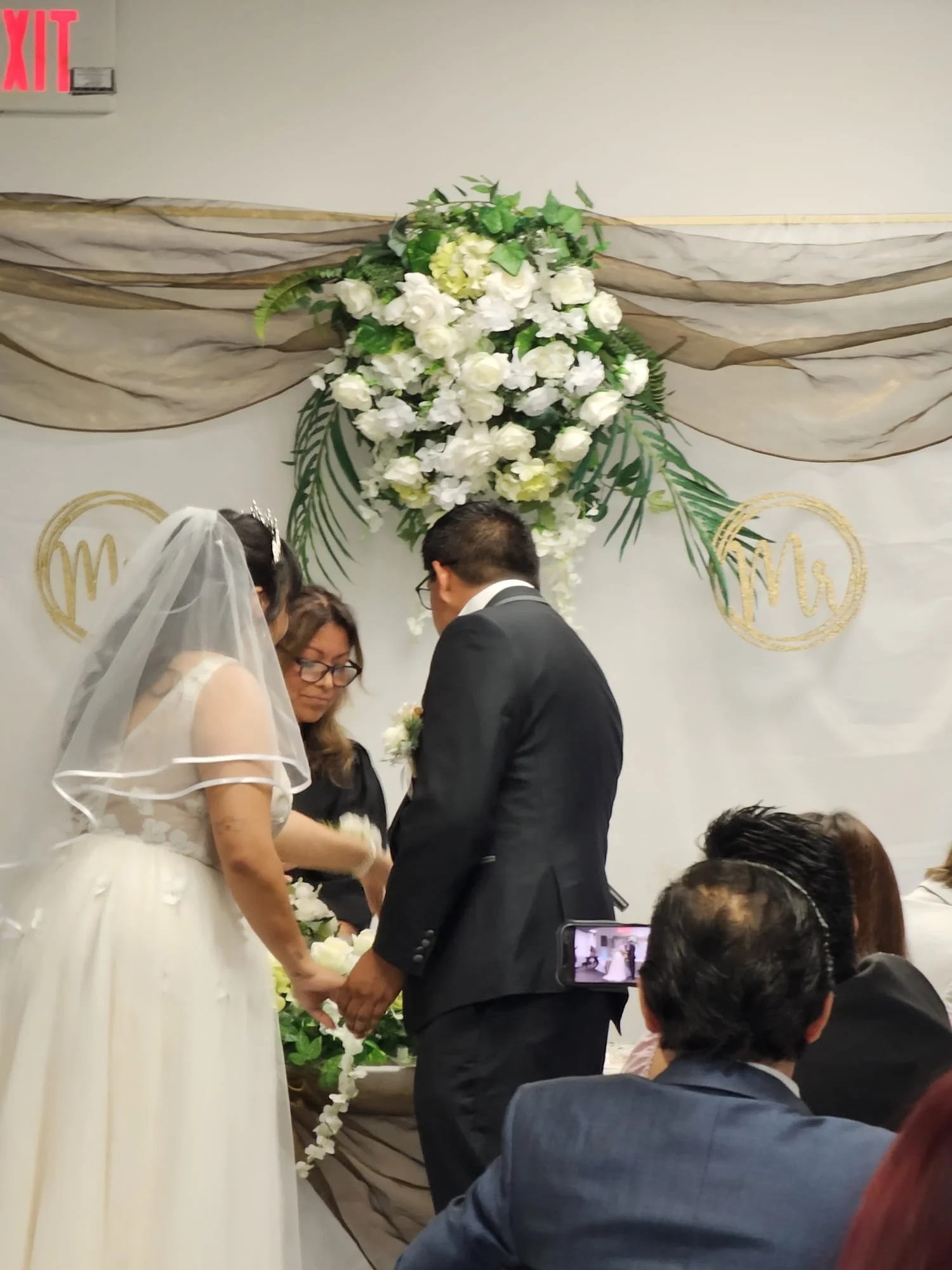Couple getting married with a wedding officiant officiating in front of floral arrangement and wedding symbols, guests seated watching.
