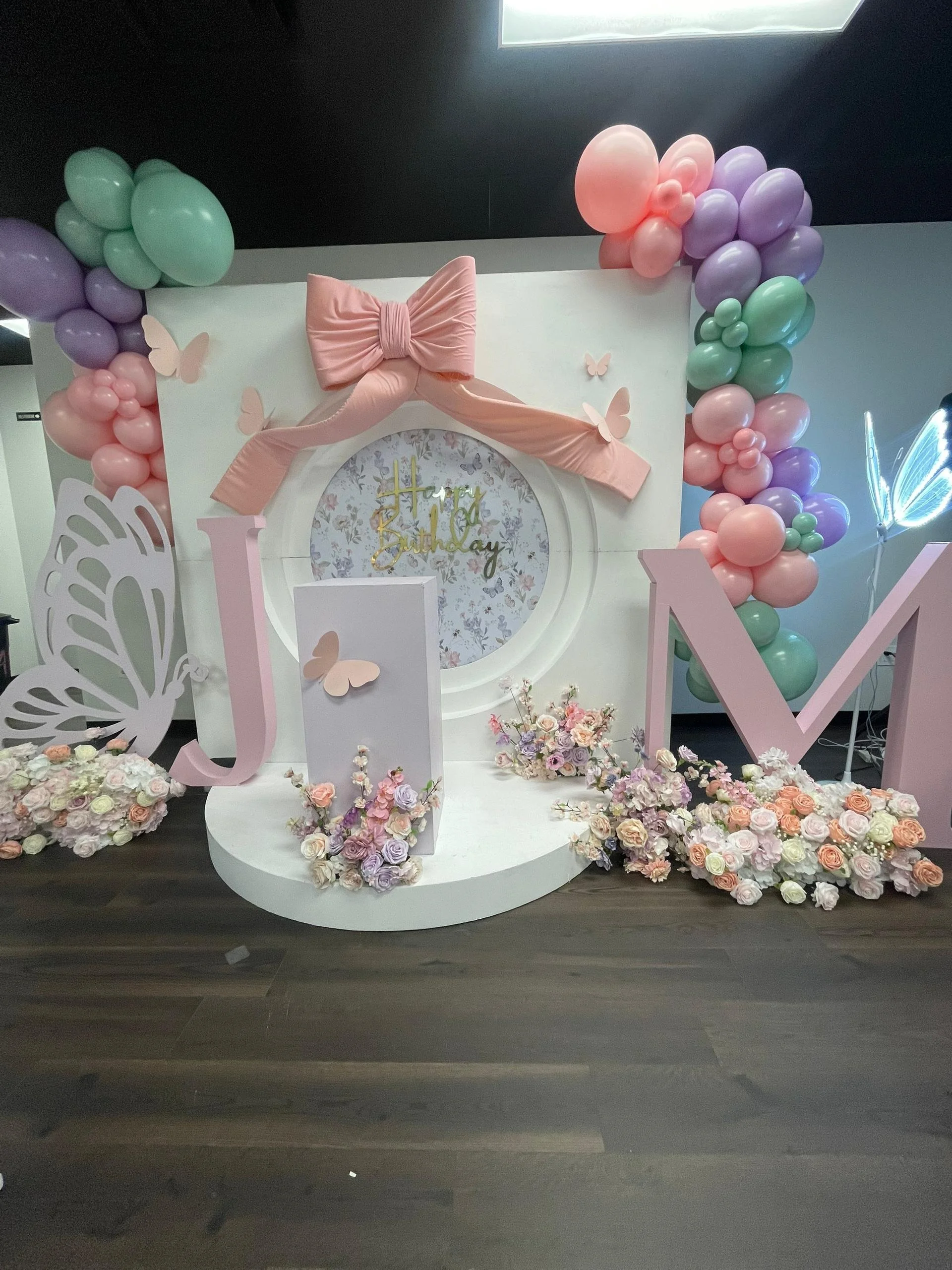 Decorative birthday party display with pink and pastel-colored balloons, floral arrangements, butterfly accents, and a 'Happy Birthday' sign.