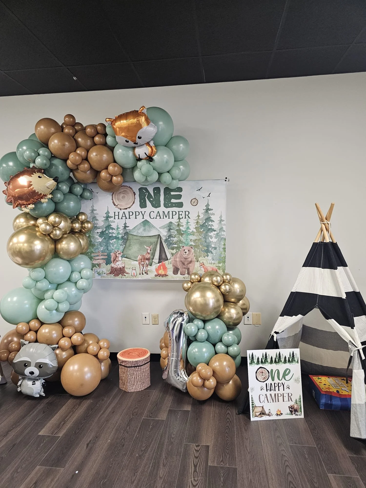 First birthday party decoration with camping theme, featuring a balloon arch with forest animal balloons, a "ONE Happy Camper" banner, and a small striped tent.