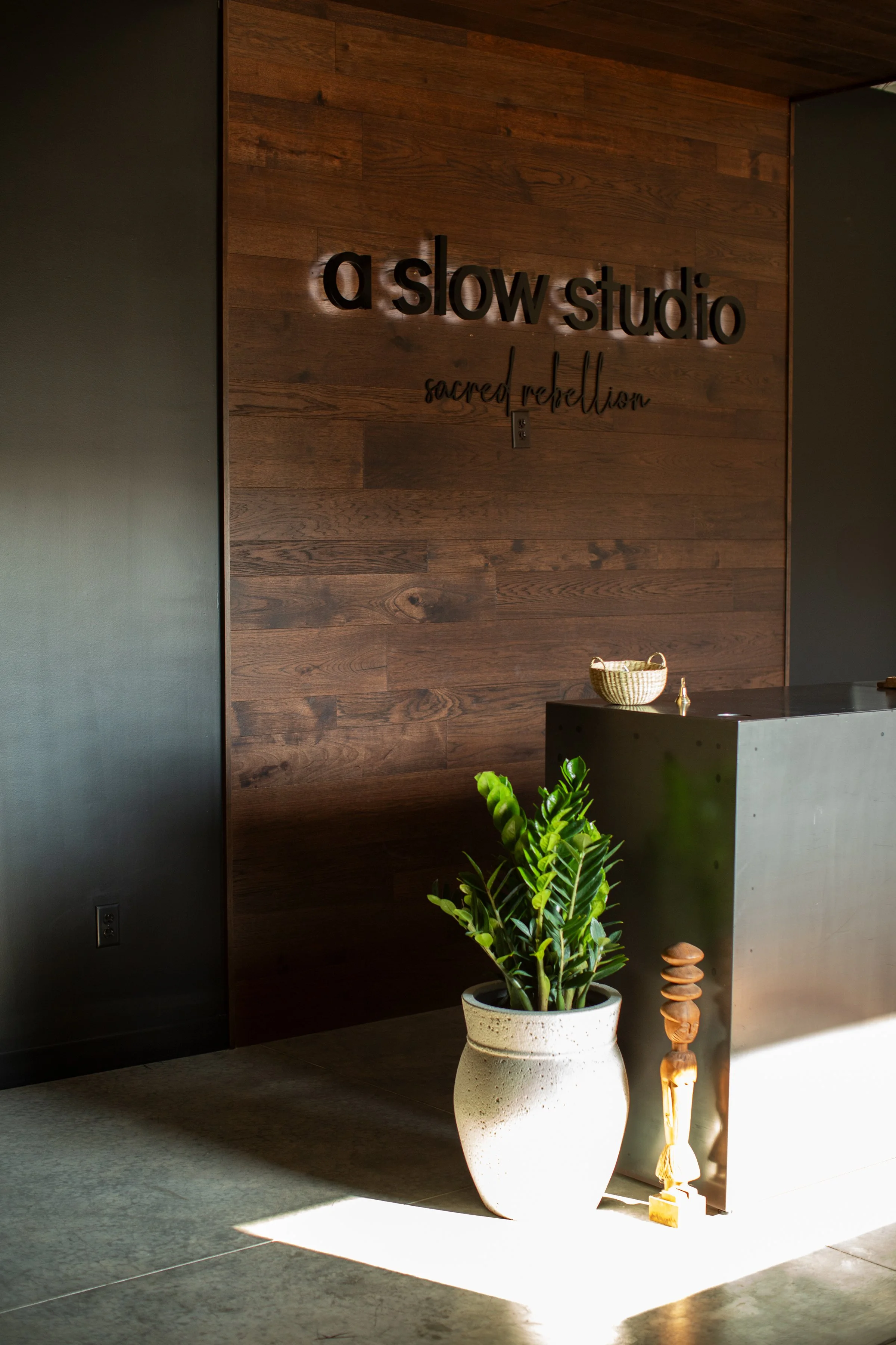 Reception desk at a studio named 'a slow studio' with the subtitle 'sacred rebellion', a potted green plant, and a small wooden sculpture next to the desk.