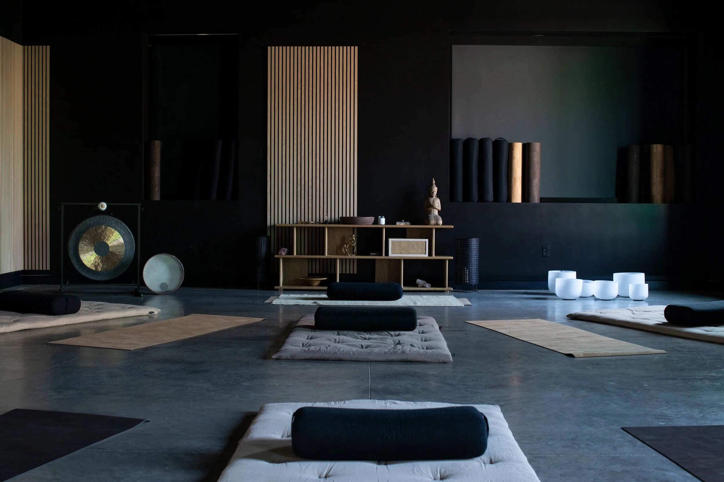 The Studio: studio with mats, bolsters, and cushions on a polished concrete floor, with wooden wall panels, a meditation gong, and a shelf with a Buddha statue and decorative objects, dimly lit.