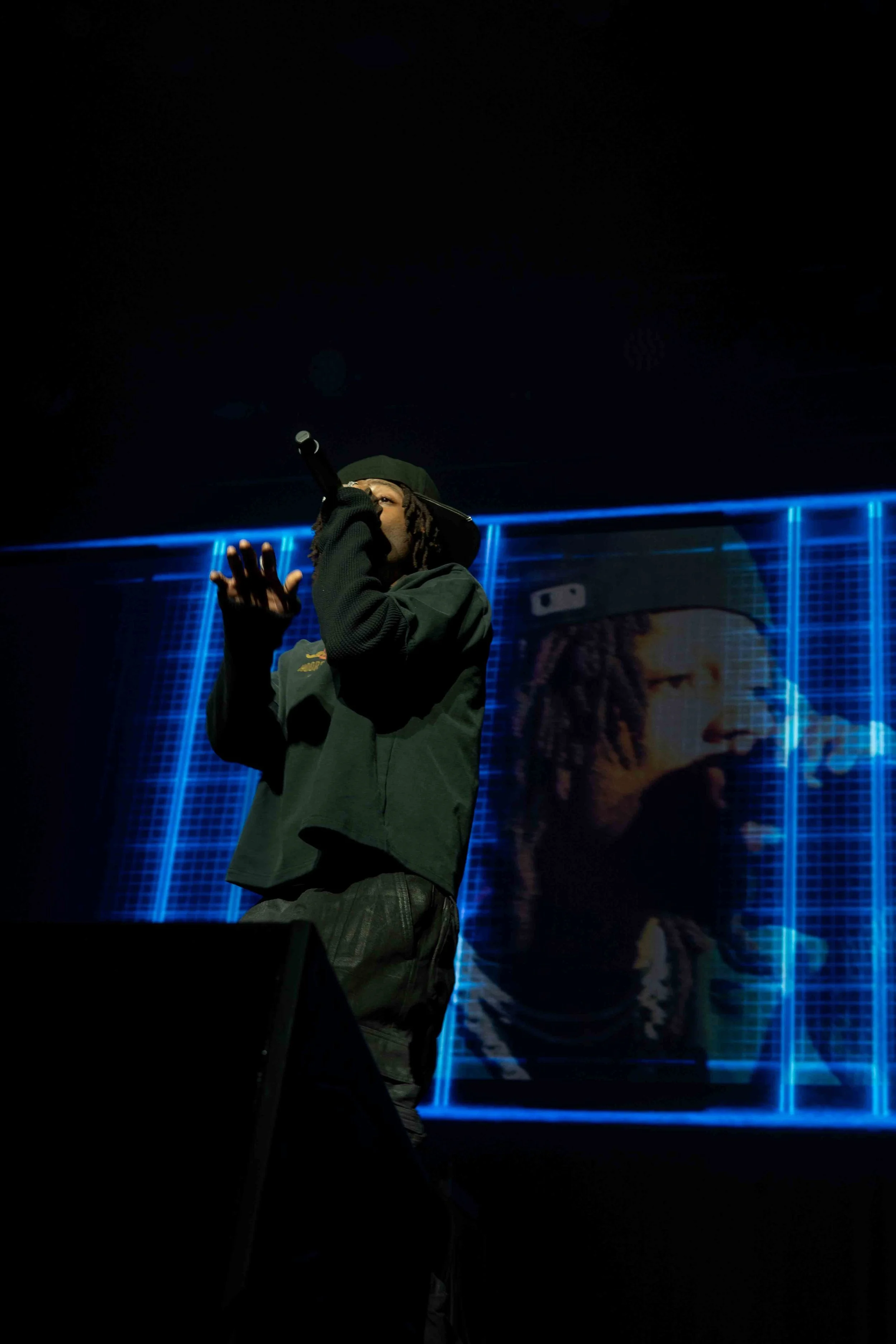 A person performing on stage, wearing a black hoodie and cap, singing into a microphone. A large digital screen in the background displays an image of the performer with a grid pattern and blue neon lights.
