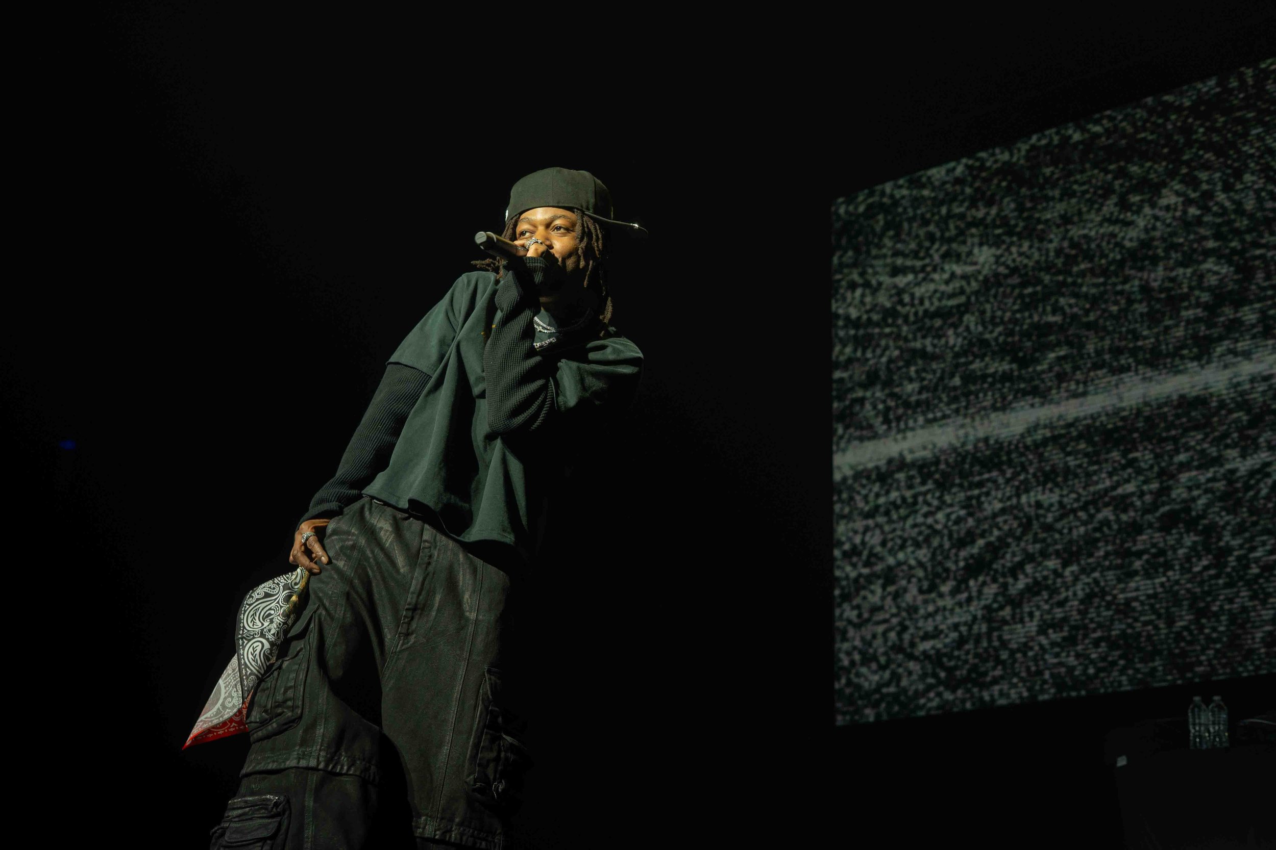 A performer with dreadlocks, wearing a black cap and dark clothing, holding a microphone and standing in front of a large screen displaying static or digital noise.