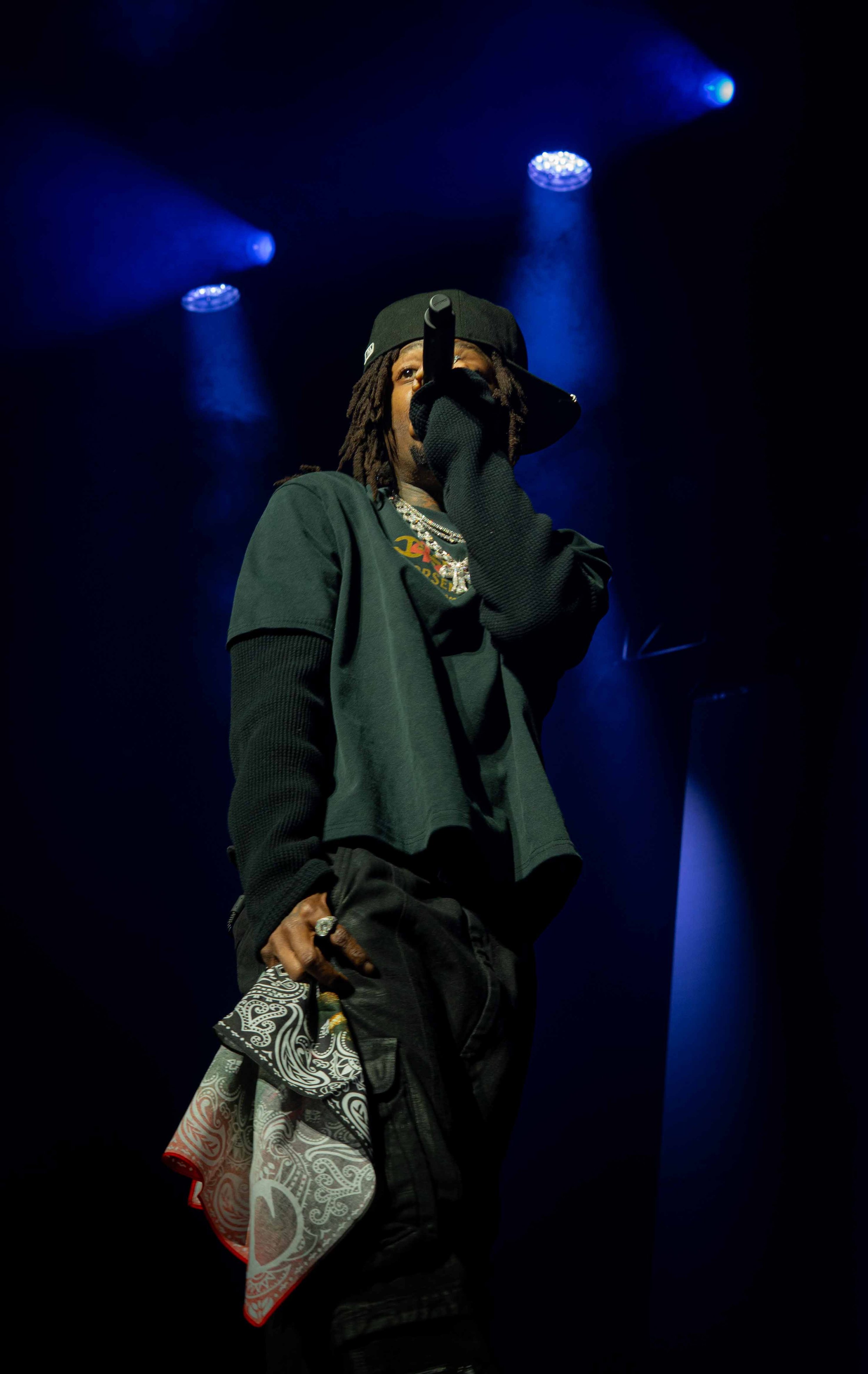 A male performer on stage holding a microphone and wearing a dark baseball cap, layered clothing, jewelry, and a patterned cloth hanging from his pants, with blue stage lights overhead.