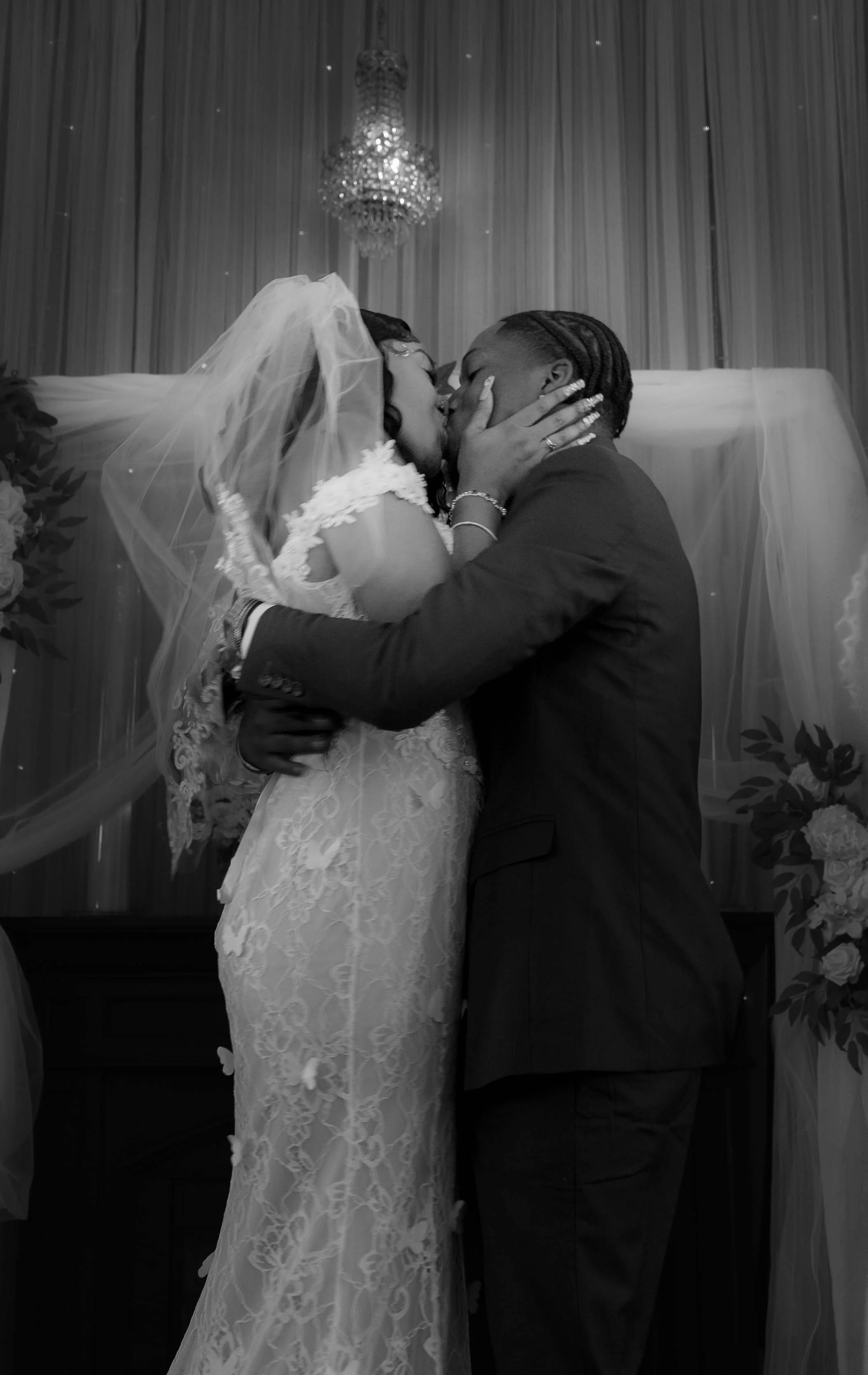 A bride and groom share a kiss during their wedding ceremony, with the bride wearing a lace wedding dress and veil, and the groom in a dark suit, standing in a decorated venue with a chandelier overhead.