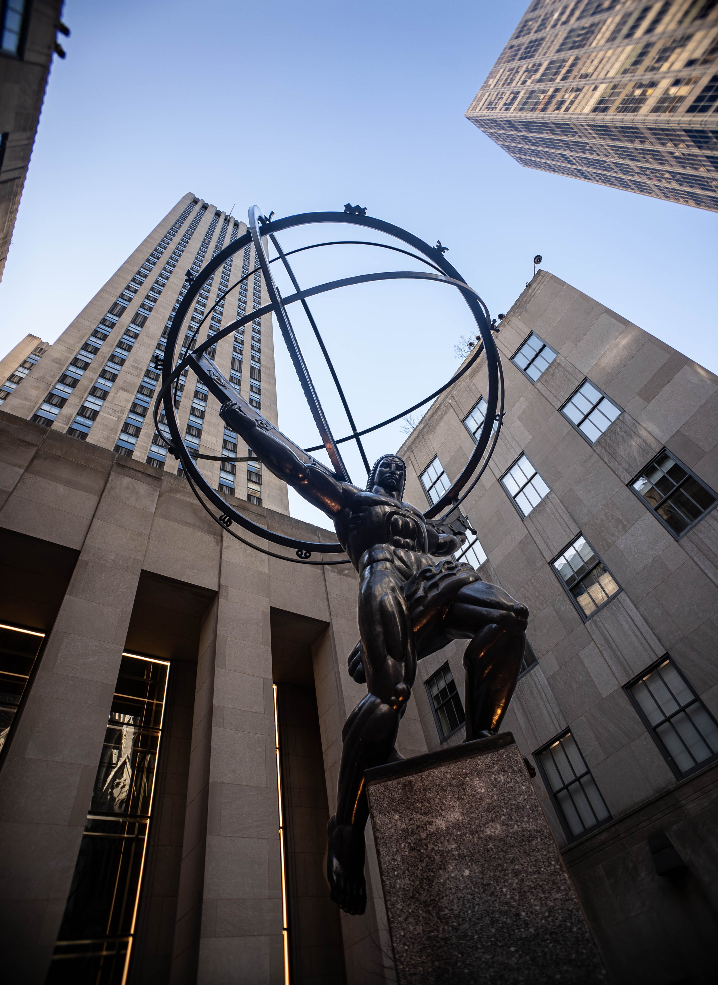 Statue of a muscular man with outstretched arm, holding what appears to be a stylized armillary sphere, set against tall skyscrapers in New York City.