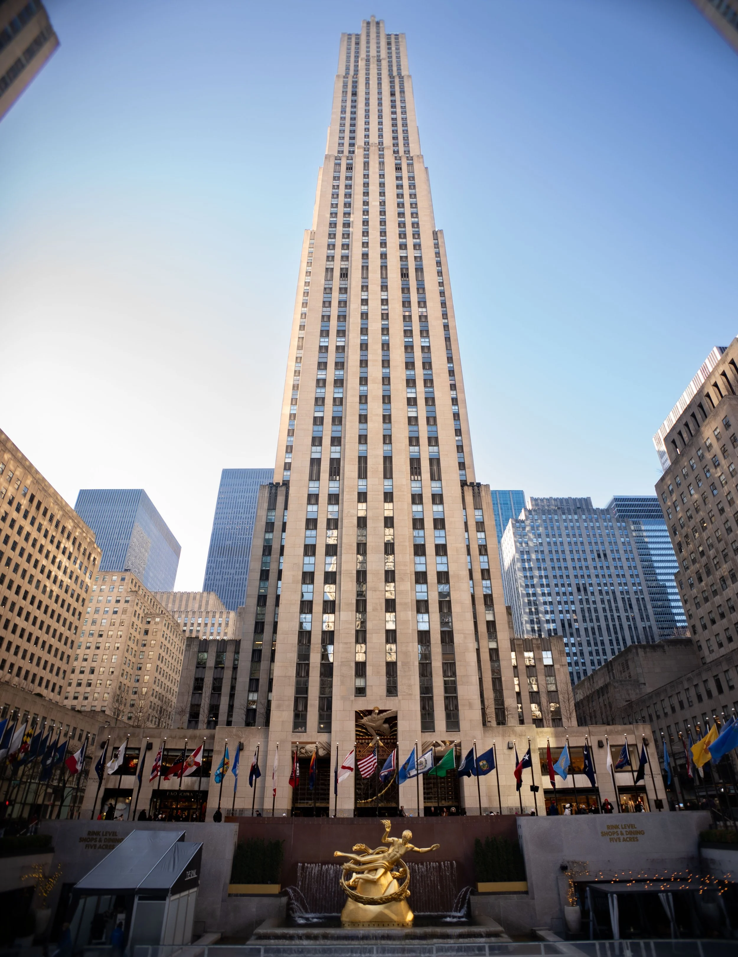 Lower view of the Rockefeller Center's GE Building in New York City with flags and a golden sculpture of a mythological figure in the foreground.