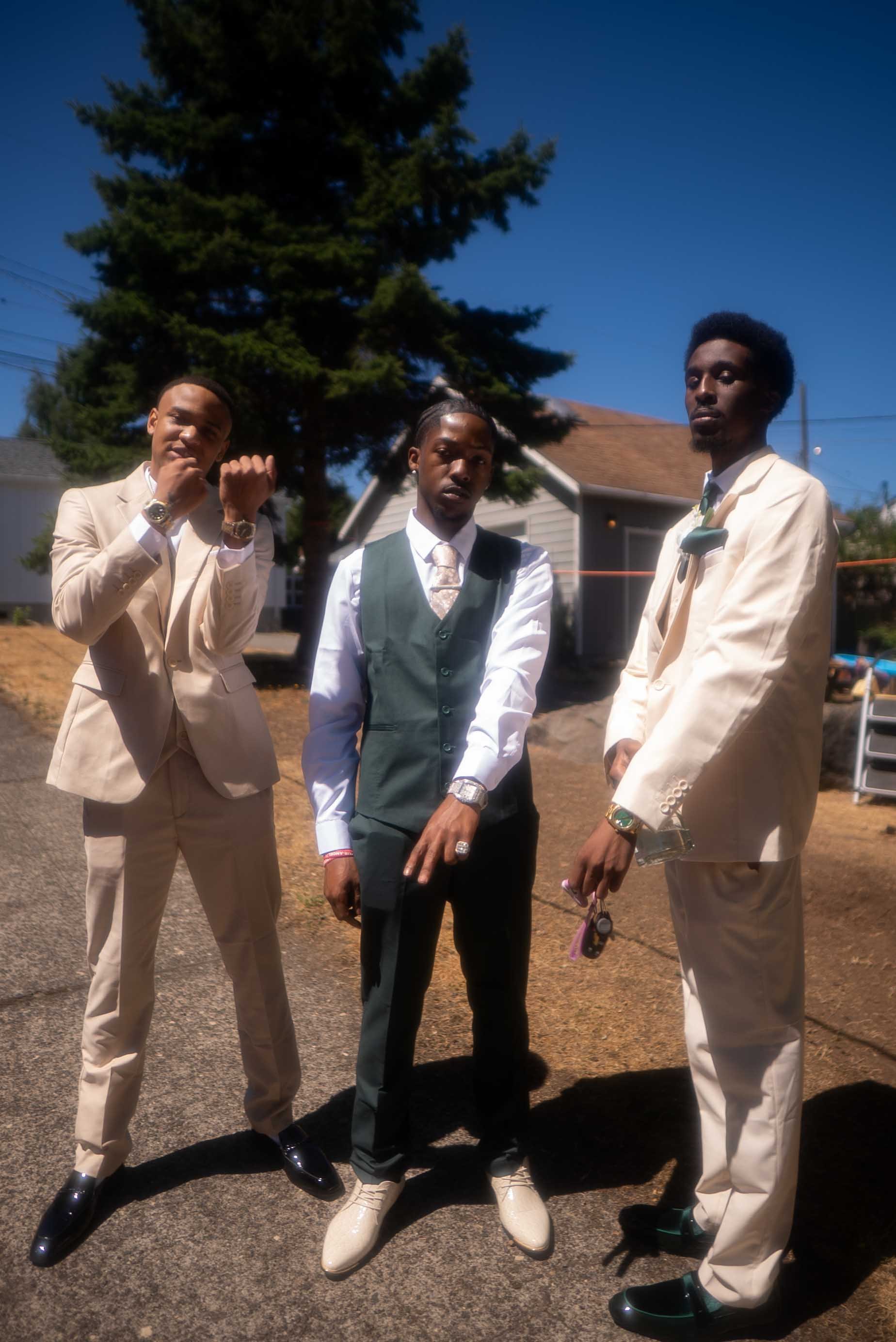 Three young men dressed in formal suits standing outdoors on a sunny day in front of a house and a large tree.