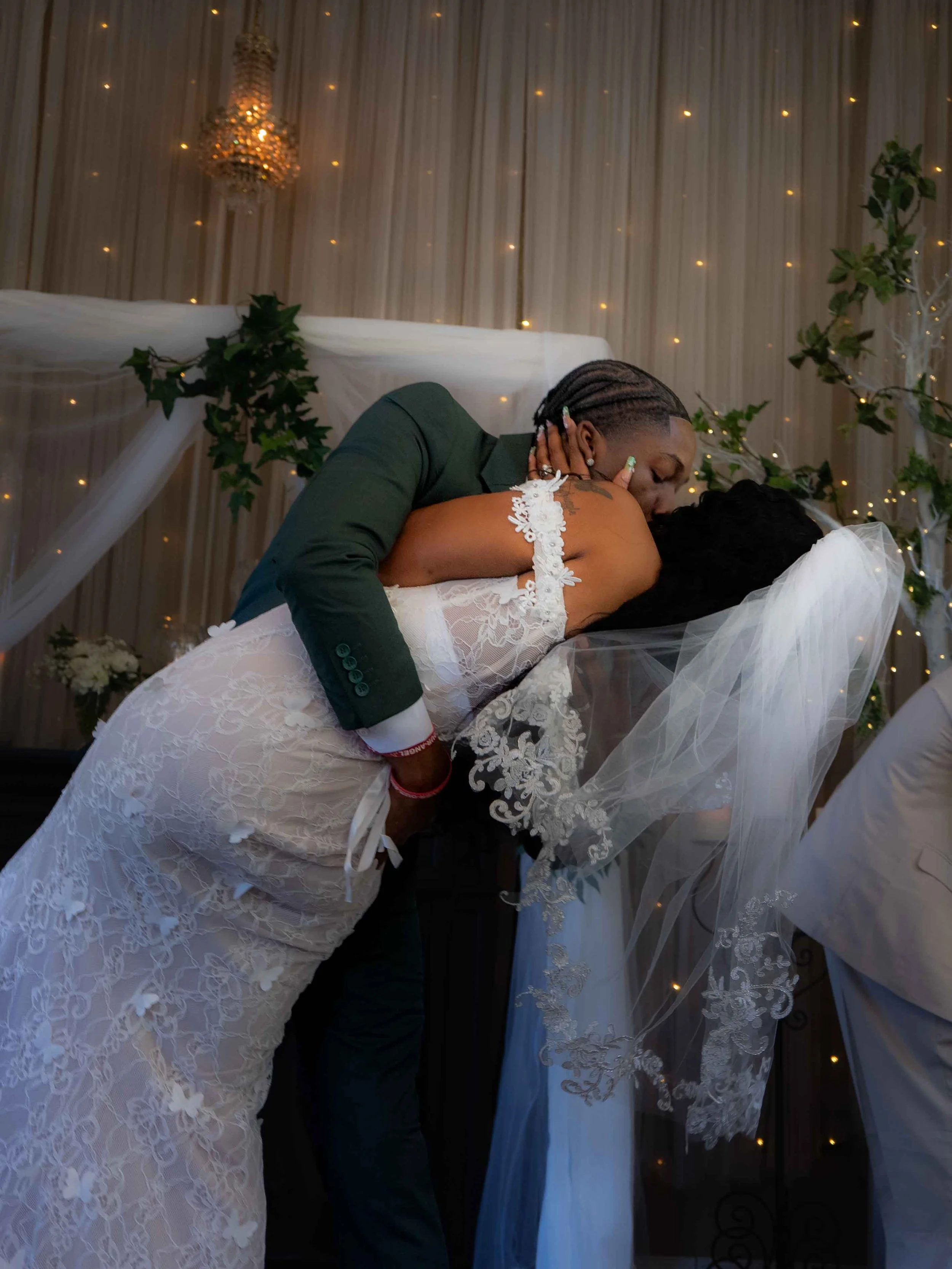 A newlywed couple sharing a kiss during their wedding reception, with the groom dipping the bride. The bride wears a lace wedding dress with a veil, and the groom is dressed in a dark green suit. The background features decorated drapes, fairy lights, and greenery.