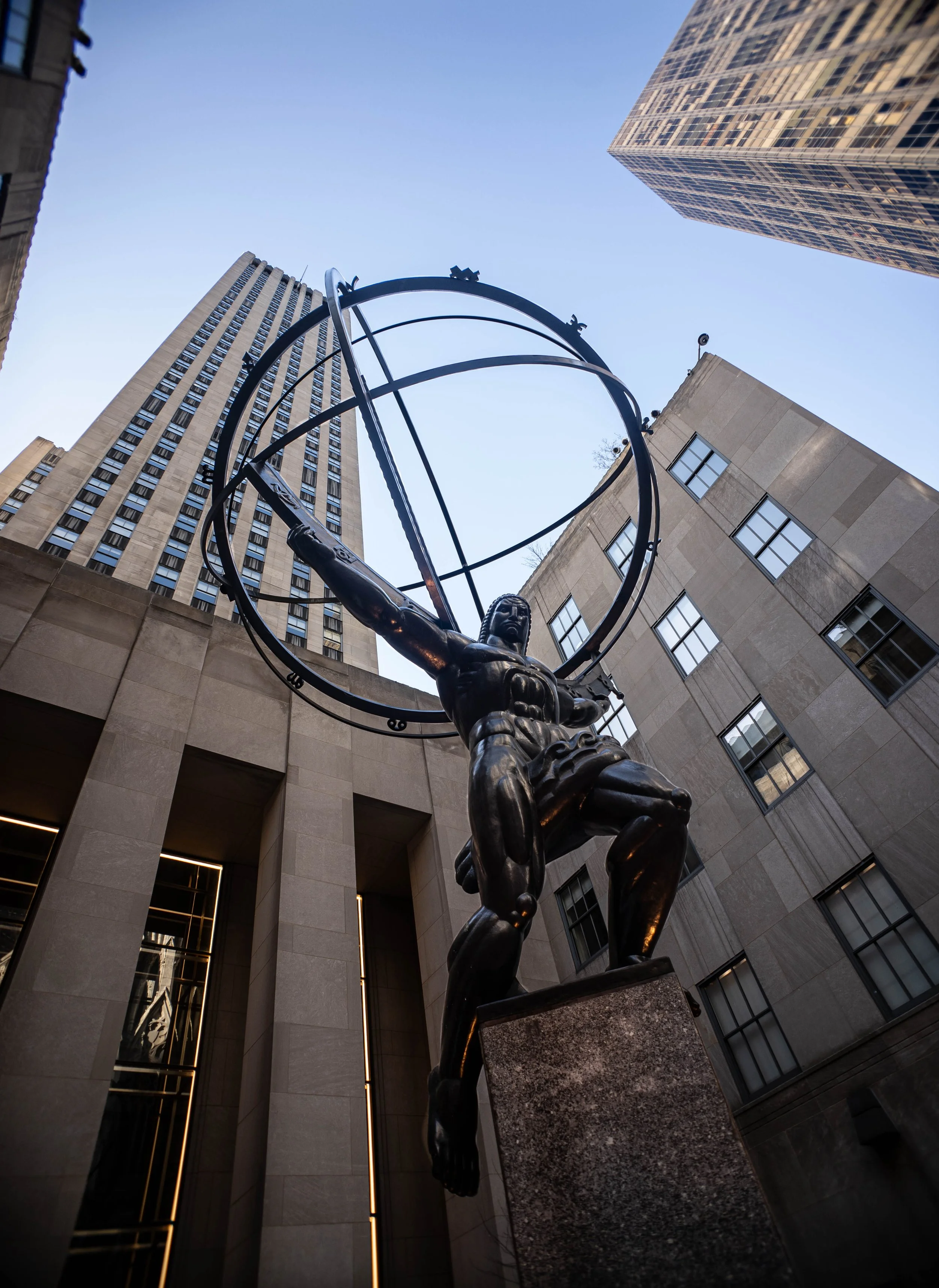 Statue of a man holding a geometric armillary sphere in an urban setting with tall buildings around.