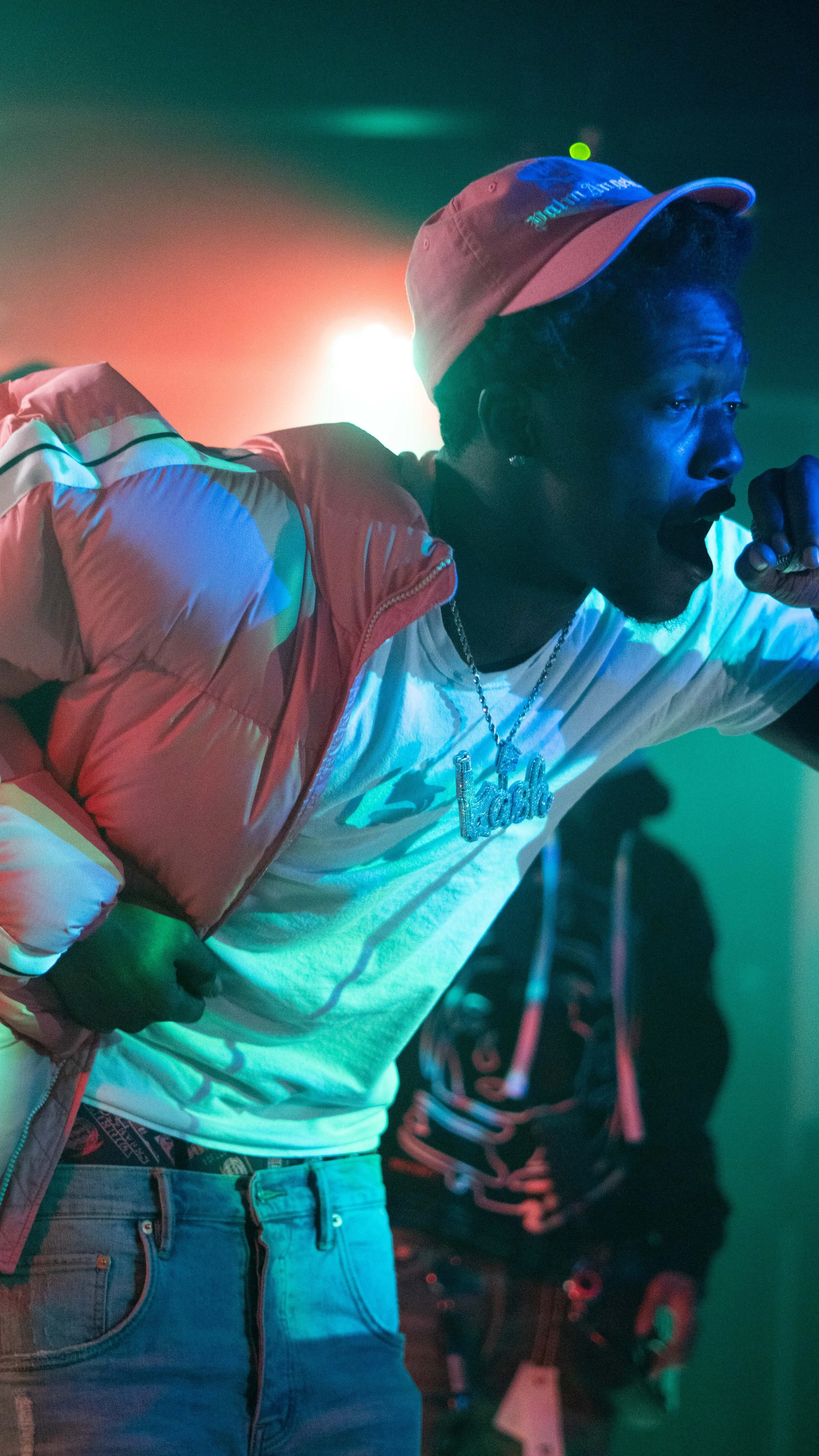 A person performing on stage holding a microphone, wearing a pink cap, white shirt, and a necklace with a large pendant; colorful stage lighting.