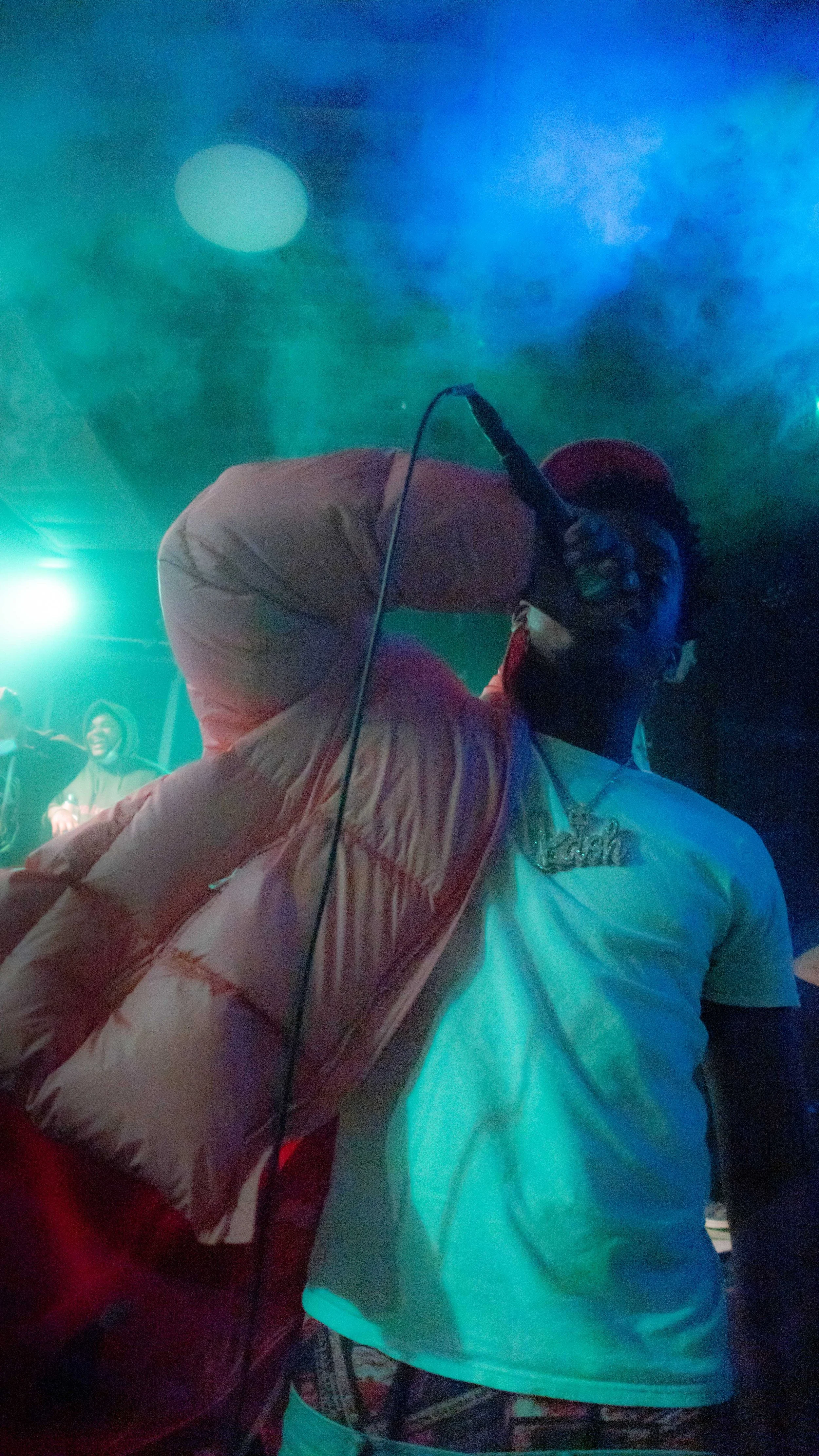 A man with long hair wearing glasses and a gold chain, holding a microphone, singing or speaking on stage with colorful lighting.