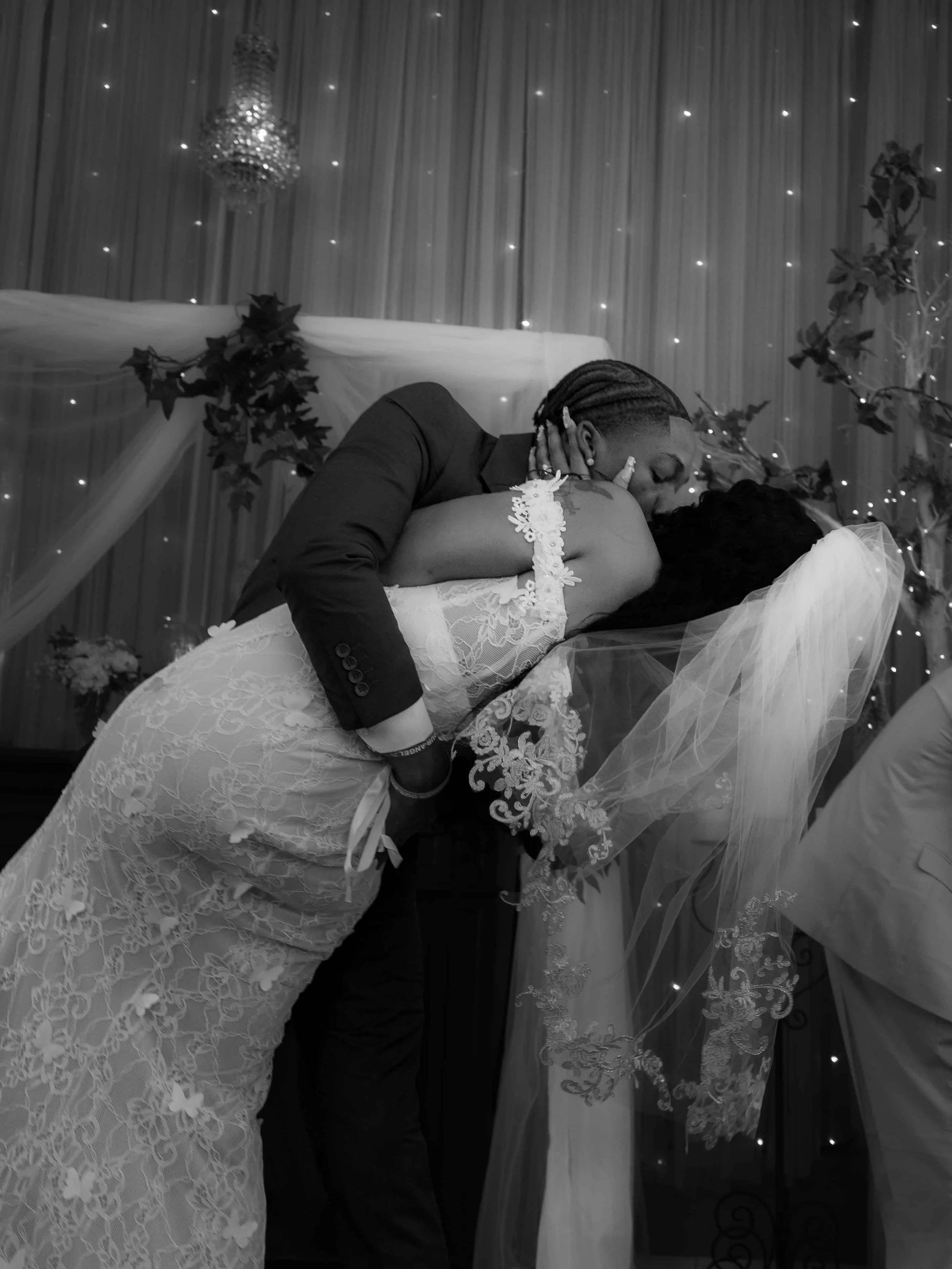 A couple in wedding attire sharing a kiss on the dance floor, with the groom leaning over the bride in a dip, amidst decorated wedding backdrop with lights and drapery.