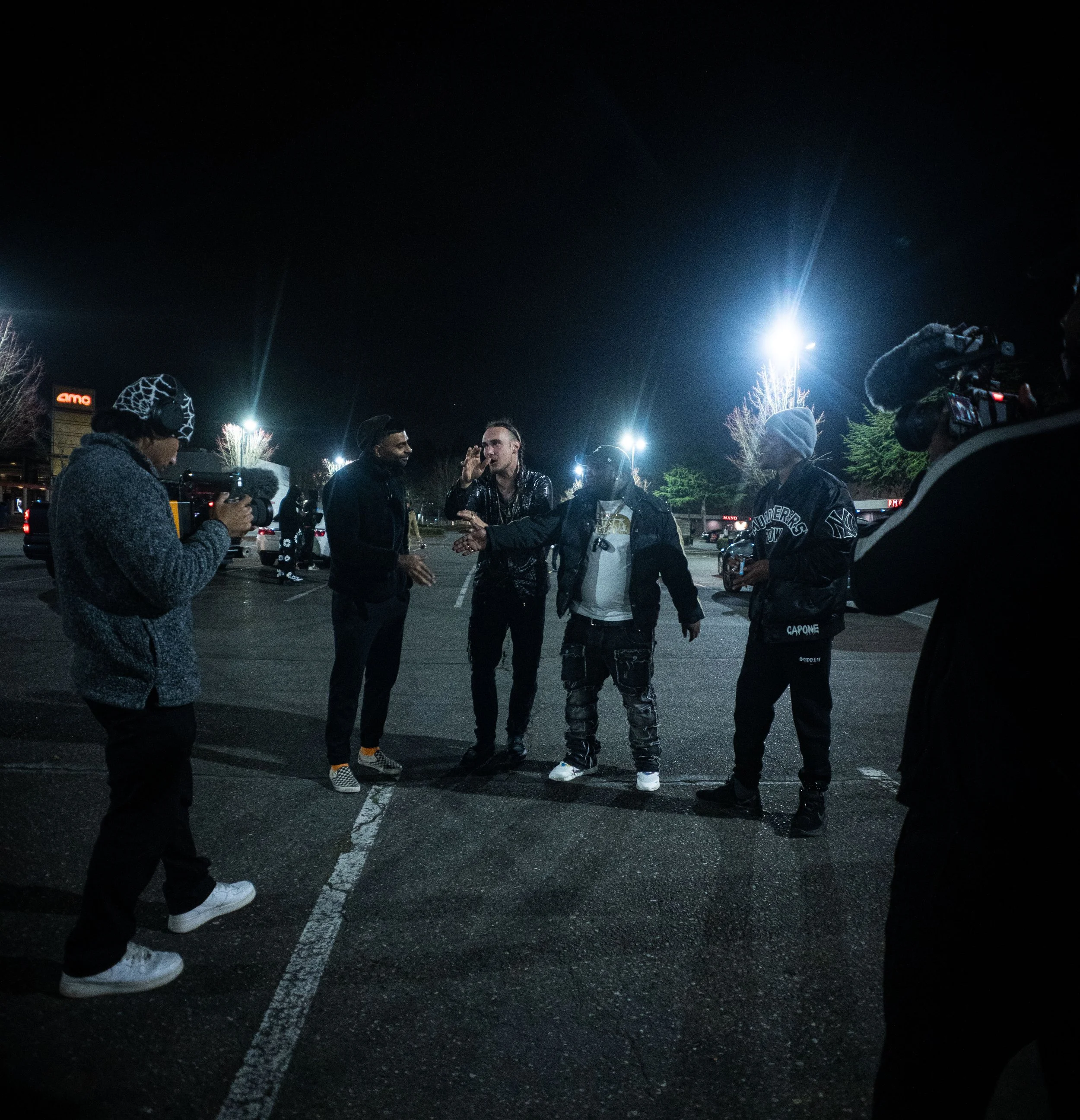 Group of people having a conversation in a parking lot at night, with one person filming and others standing around, illuminated by bright overhead lights.