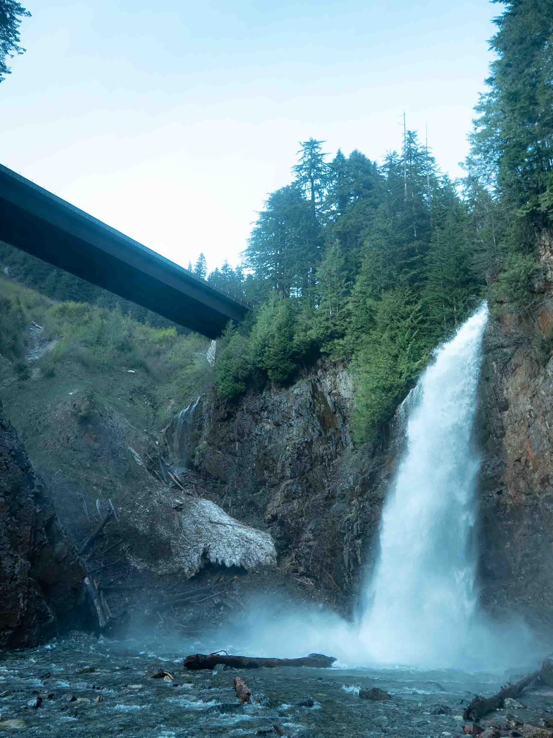 Tall waterfall cascading down a rocky cliff into a river, surrounded by lush green trees, with a bridge arching overhead.
