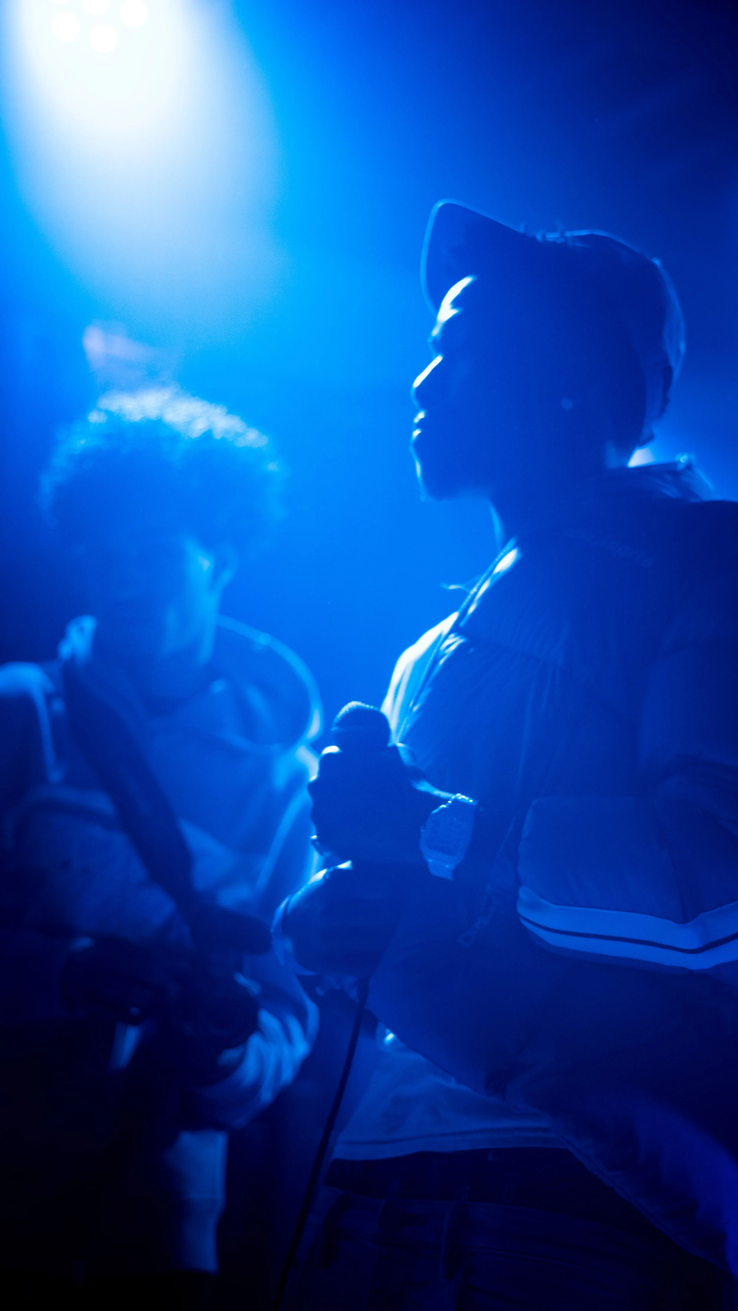 Silhouettes of two people in a dark, blue-lit environment, one wearing a cap and the other wearing glasses, engaging in a conversation.