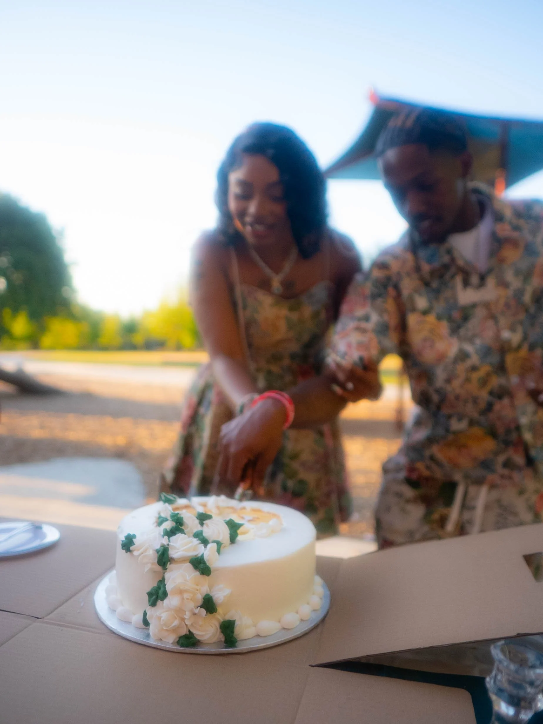 A couple cutting a white wedding cake decorated with green leaves and white flowers at an outdoor celebration under a canopy, with trees and a clear sky in the background.
