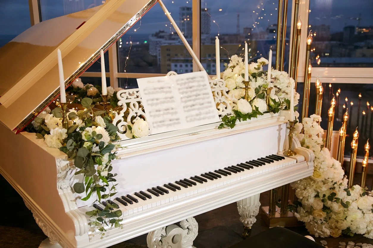 White grand piano decorated with white flowers, greenery, and lit candles, set in a room with city view windows at dusk.