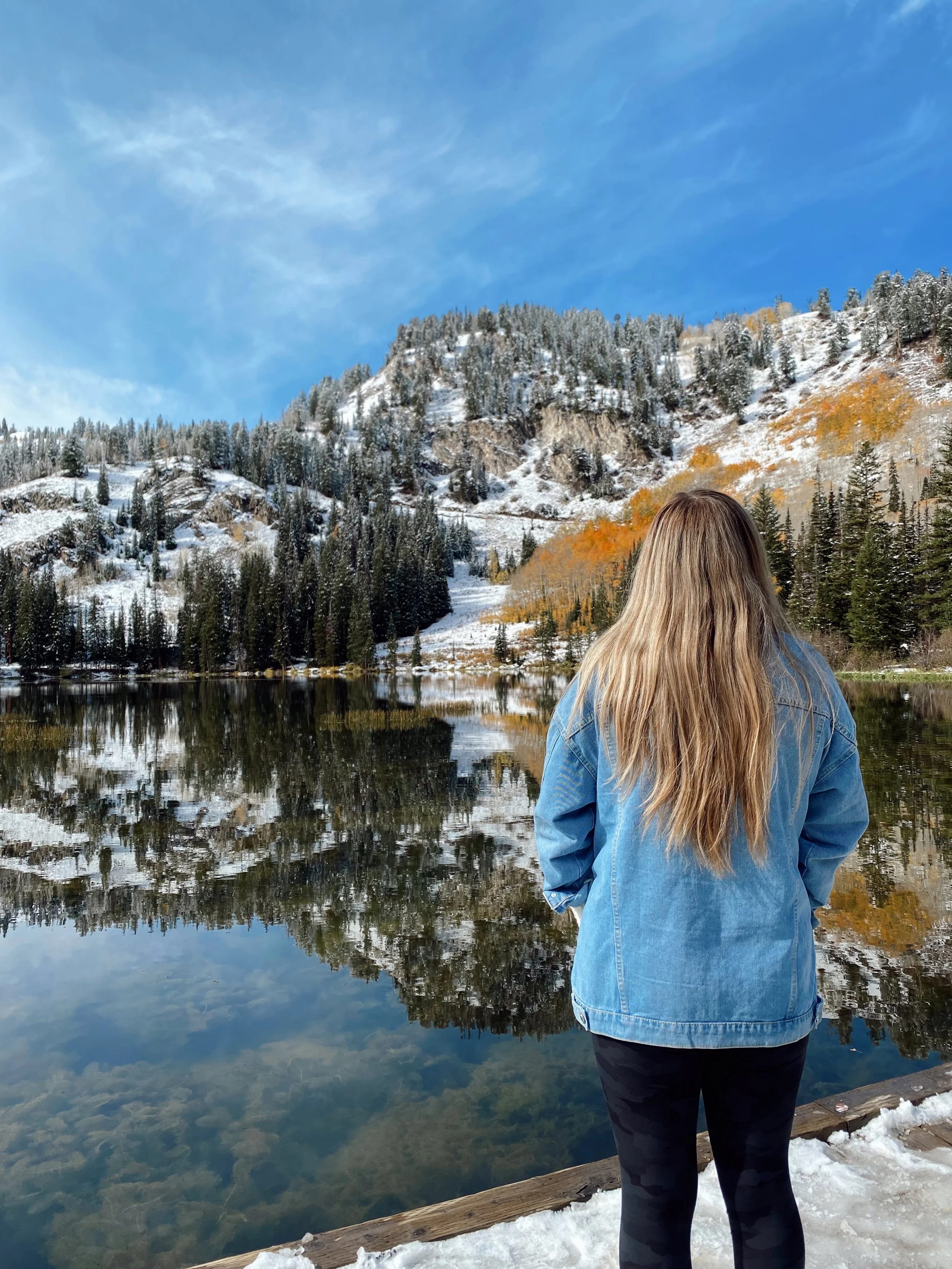 A woman with long blonde hair wearing a blue denim jacket and black pants standing on a snow-covered wooden dock, looking at a calm lake with a mountain and forest in the background.
