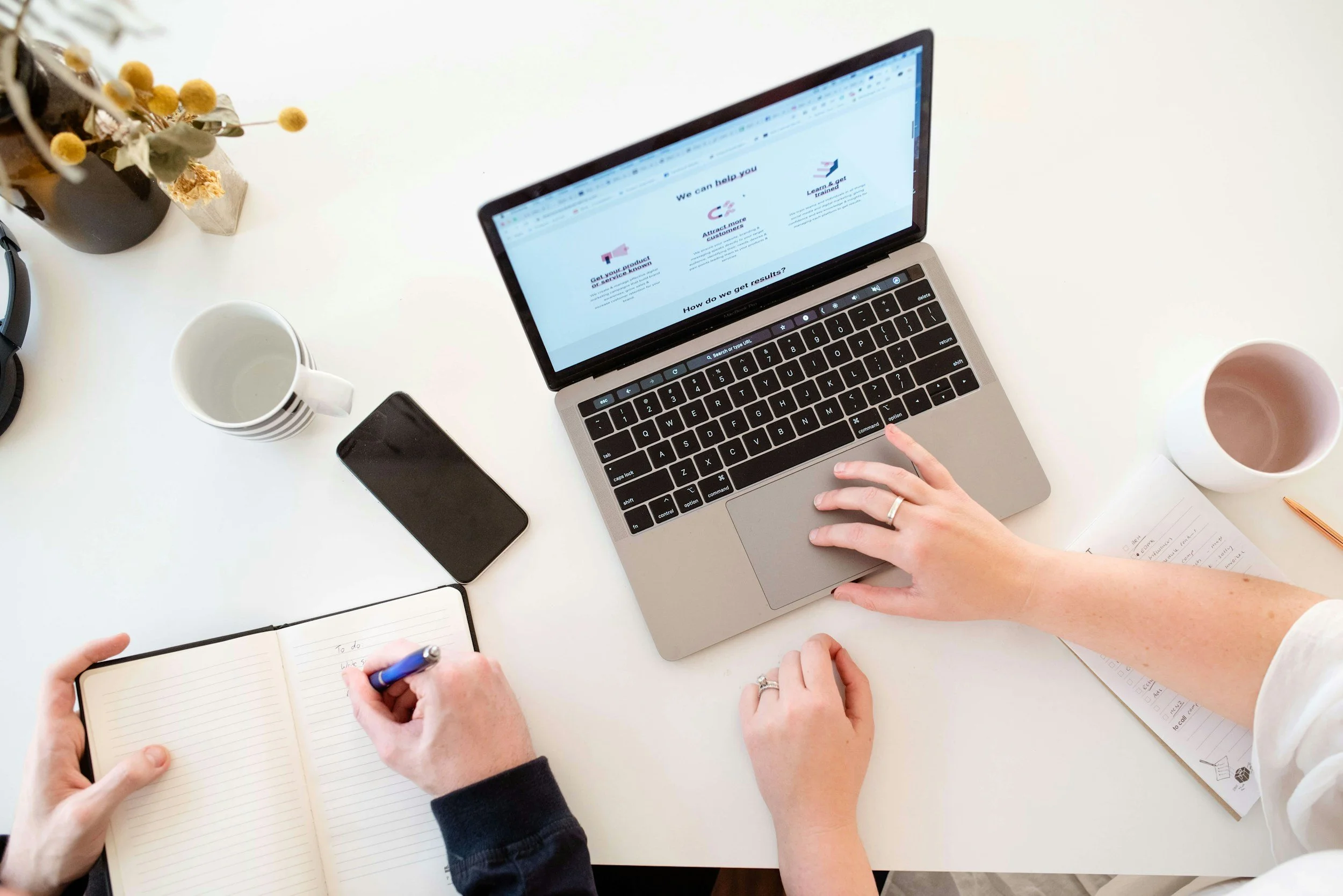 Top-down view of two people working at a white desk with a laptop, smartphone, notebooks, mugs, and a plant.