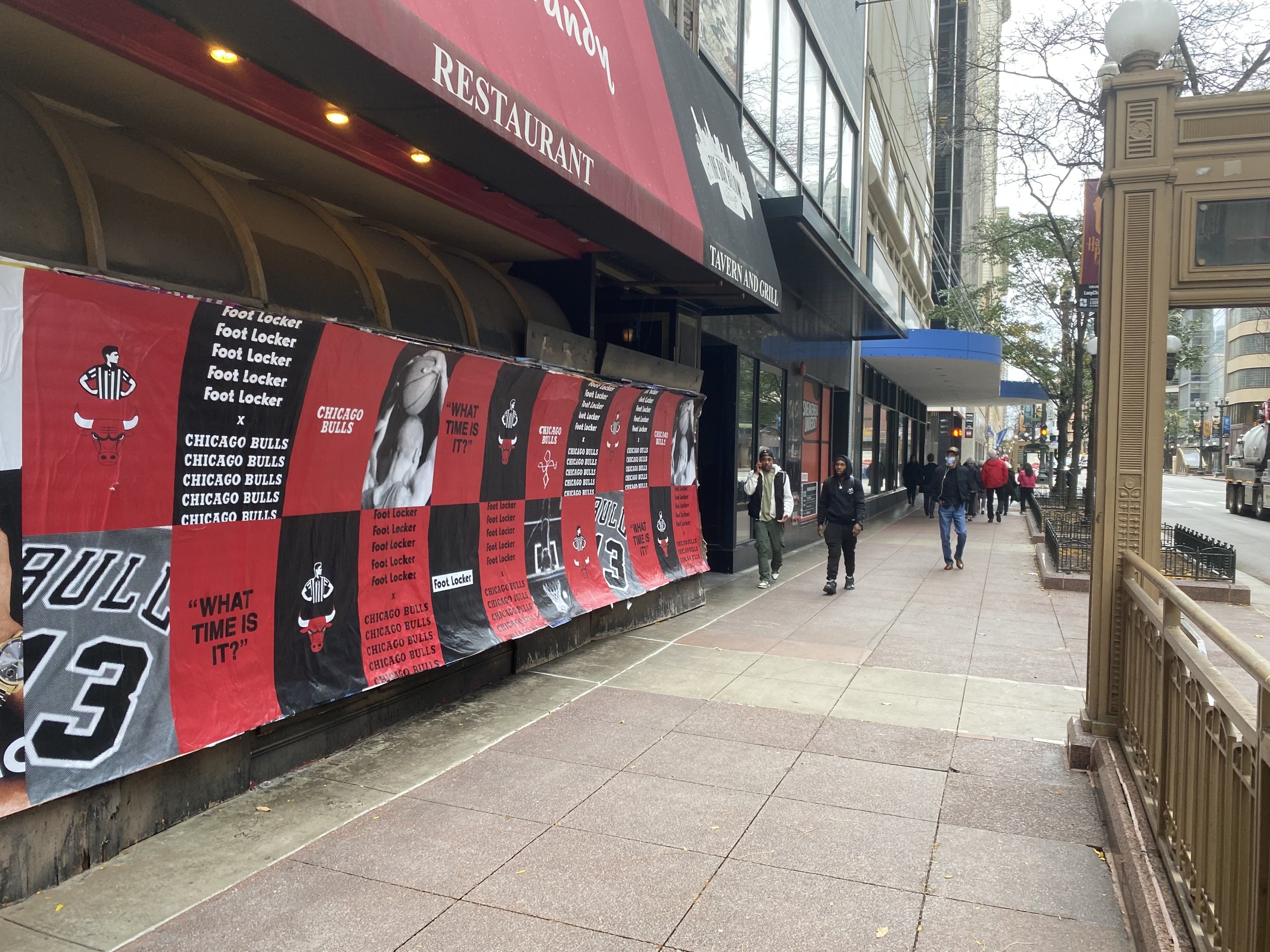 City sidewalk with pedestrians walking past a restaurant with red advertising banners on the wall featuring images of Chicago Bulls basketball team and the slogan "What time is it?" in black and red. Some trees and street lights are visible in the ba