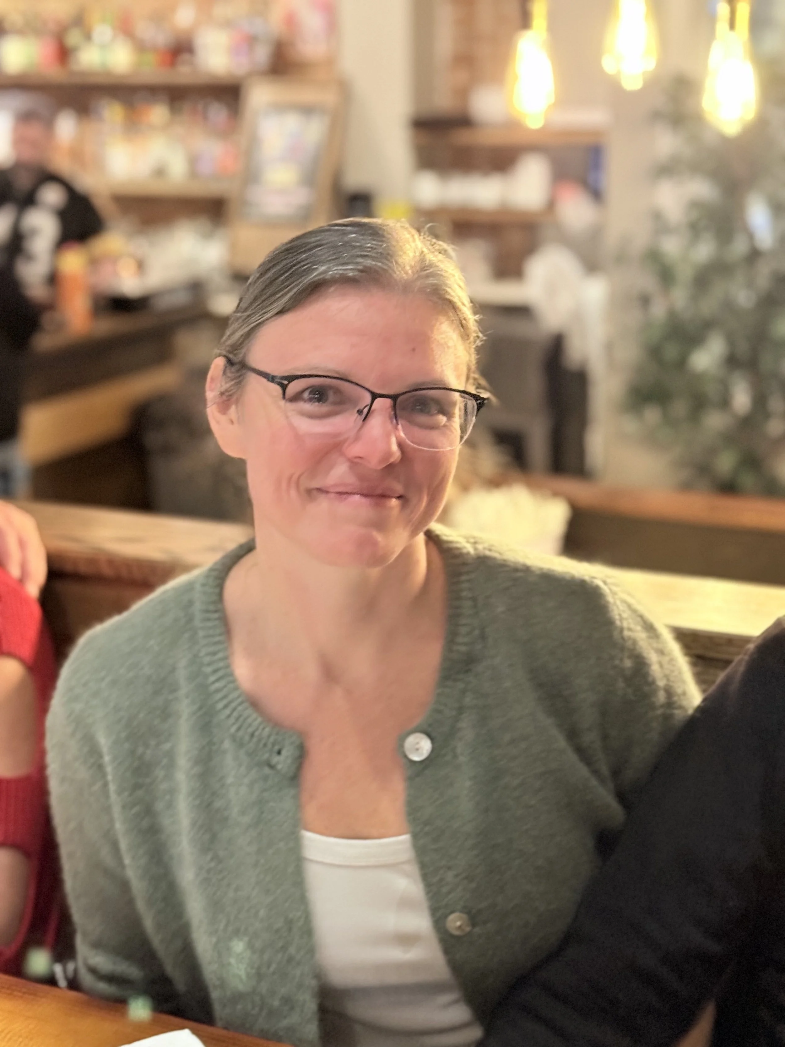 A woman with short gray hair and glasses, smiling, seated in a cozy restaurant or cafe with warm lighting and bookshelves in the background.