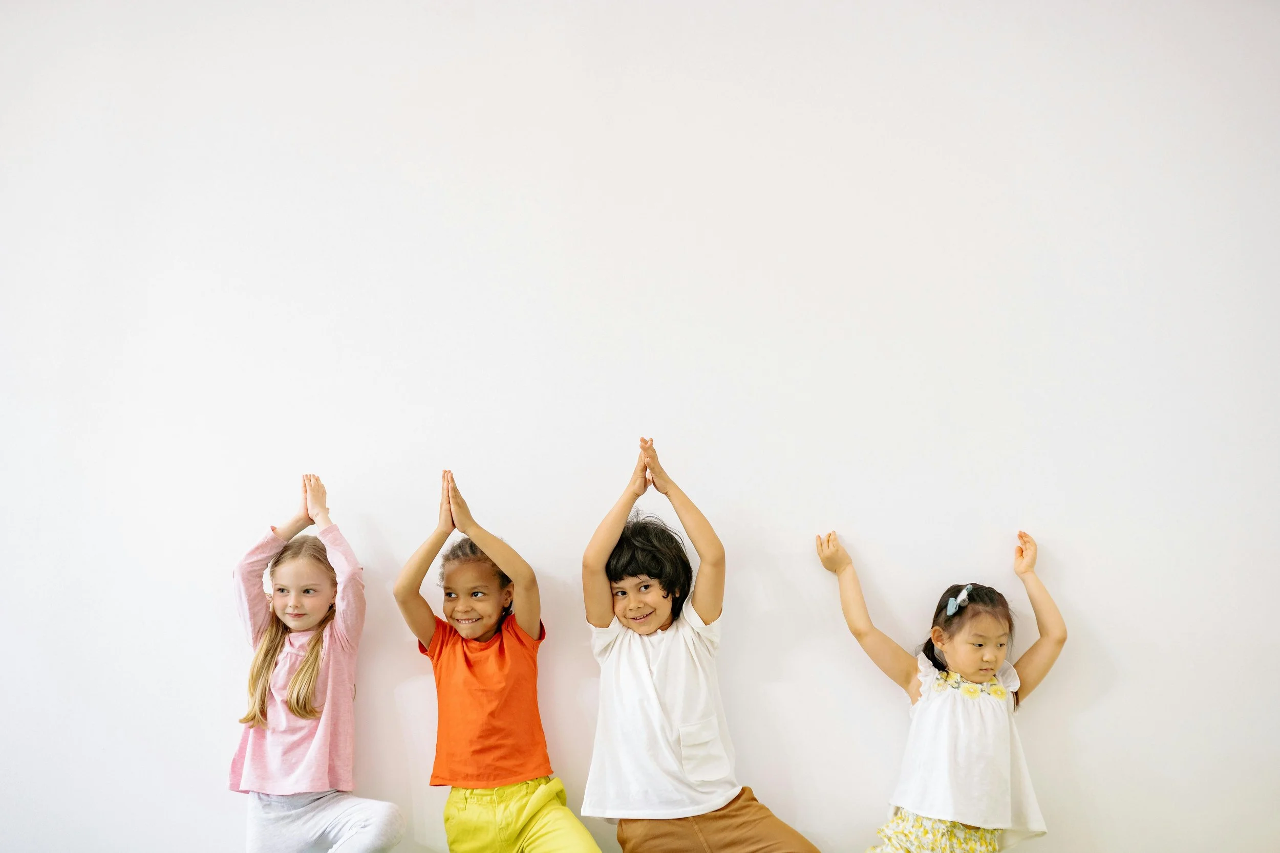 Four children standing against a plain white wall, with arms raised and hands pressed together above their heads, smiling and posing playfully.