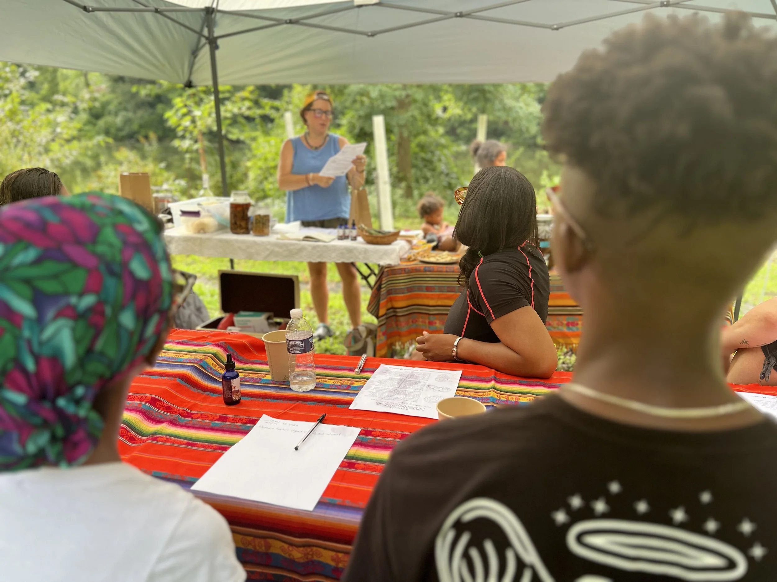 People sitting around a colorful table under a canopy, listening to a woman speaking at a table with various items, in an outdoor setting.