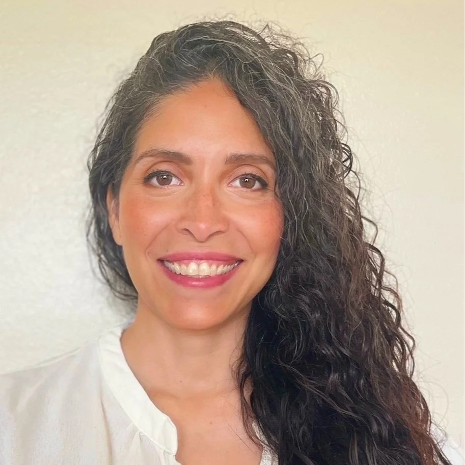 A woman with curly dark hair smiling at the camera over a plain light-colored background.