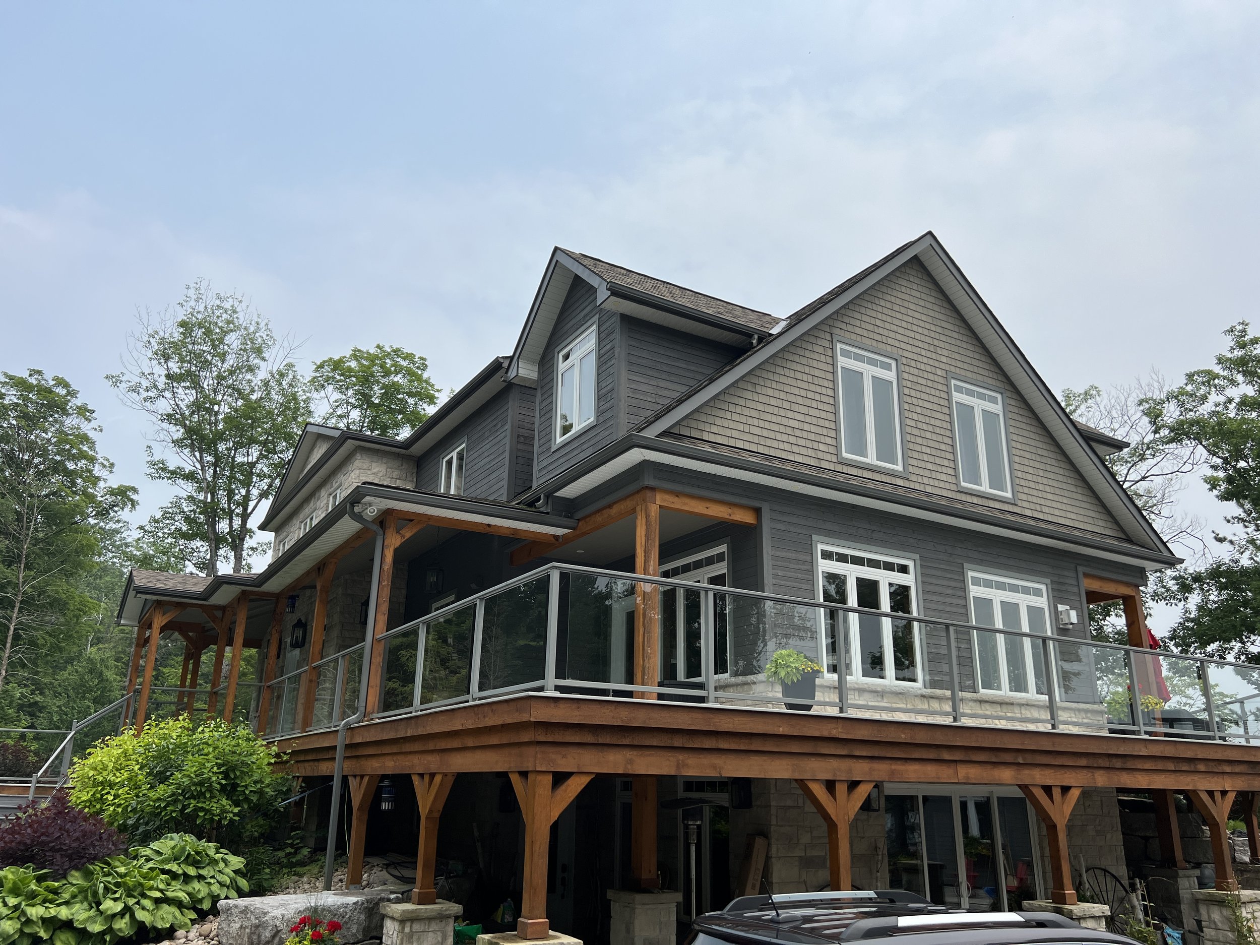 Two-story gray house with wooden accents and a large deck, surrounded by trees.