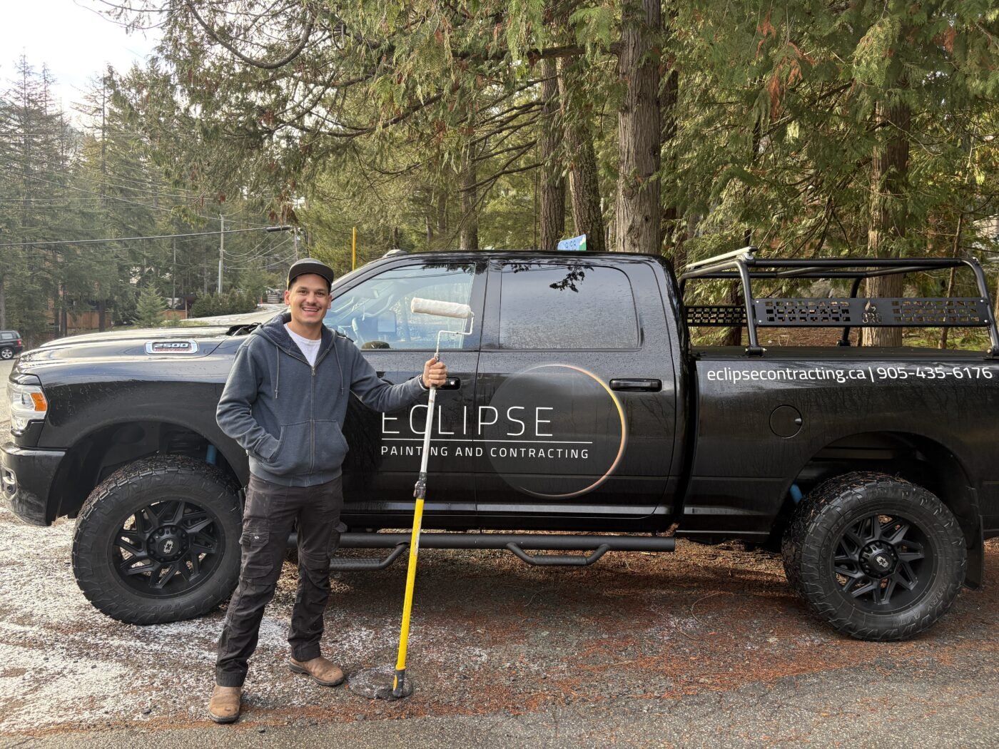 A smiling man in a gray hoodie, black pants, and a cap holding a paint roller stands next to a black pickup truck branded with Eclipse Painting and Contracting. The truck is parked outdoors with trees in the background.