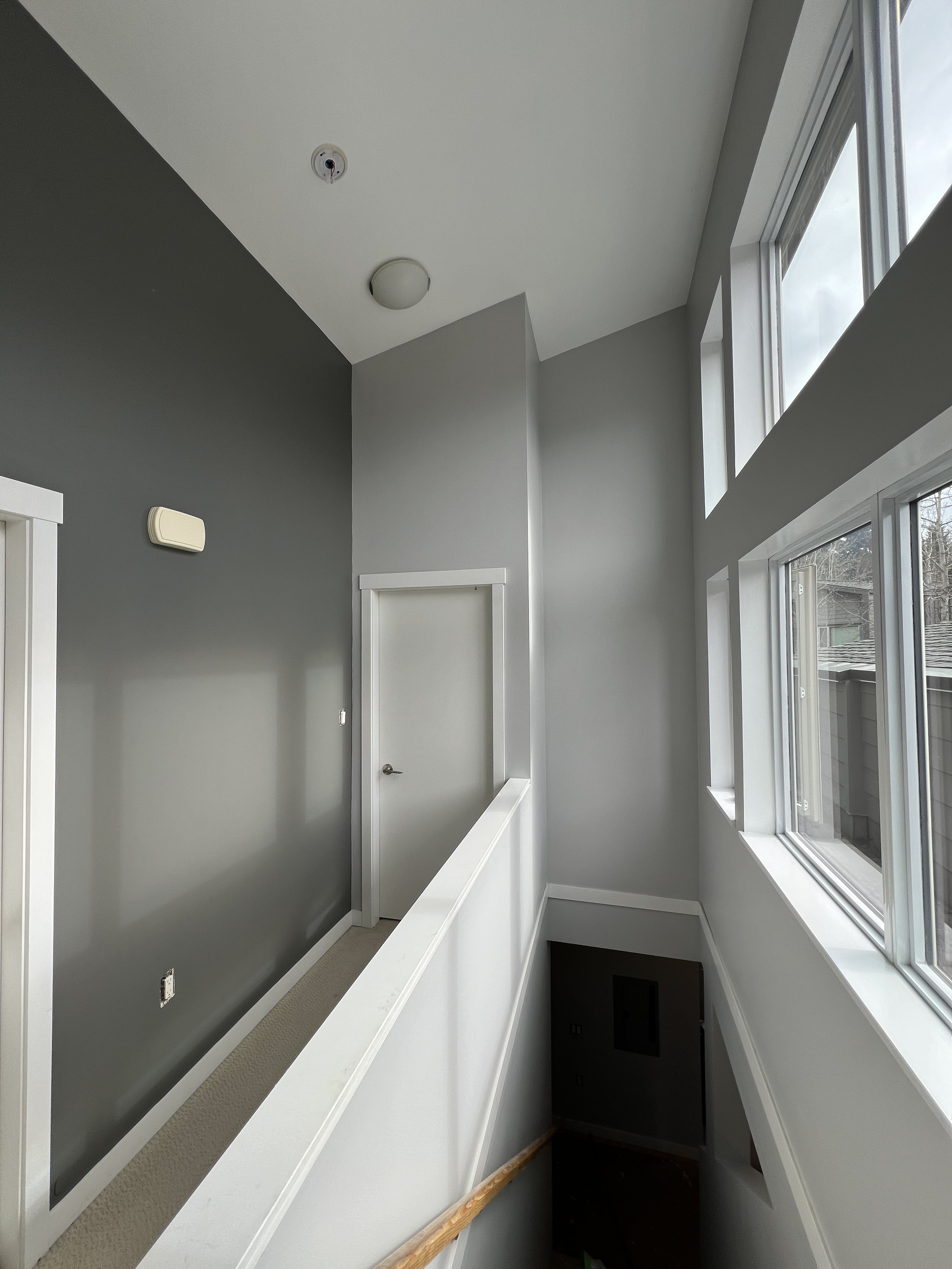 Interior view of a house with gray and white walls, large windows, and a hallway leading to a closed door. Ceiling has a light fixture and smoke detector. The staircase is partially visible below.