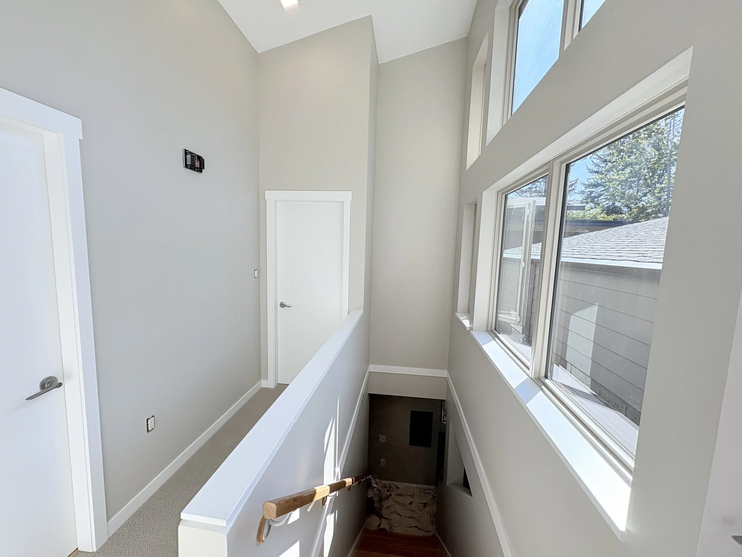 A bright, modern interior with large multipane windows, white walls, and a stairway leading down, with a door at the top of an upper hall.