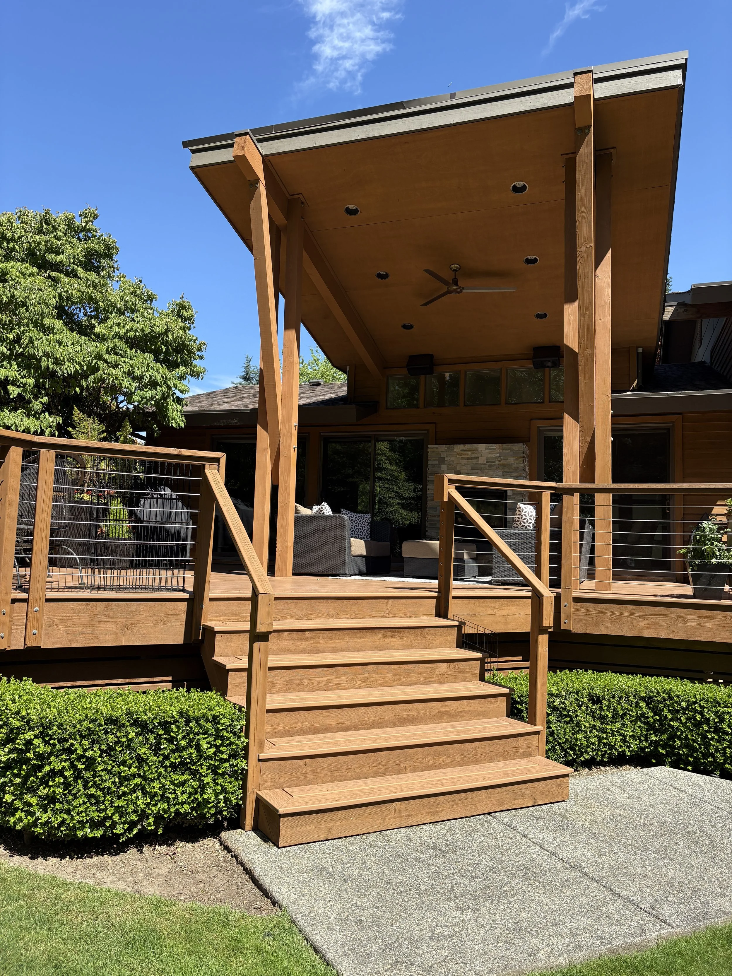 Backyard wooden patio with steps, covered by a roof, outdoor seating area, and green landscaping.
