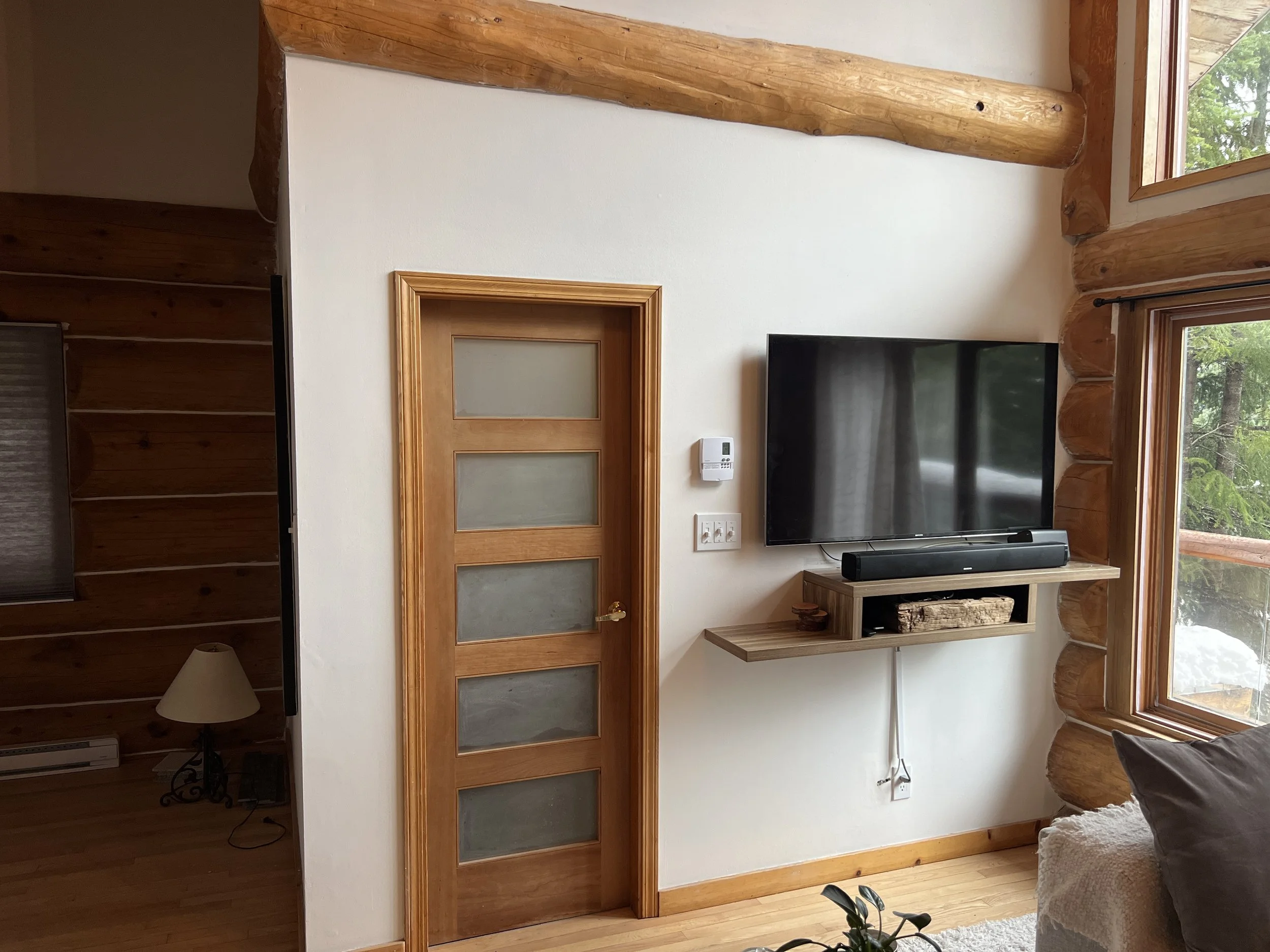 Interior of a cozy wood cabin living room with a flat-screen TV mounted on a white wall, wooden shelves, a window with nature view, and a door with glass panels.