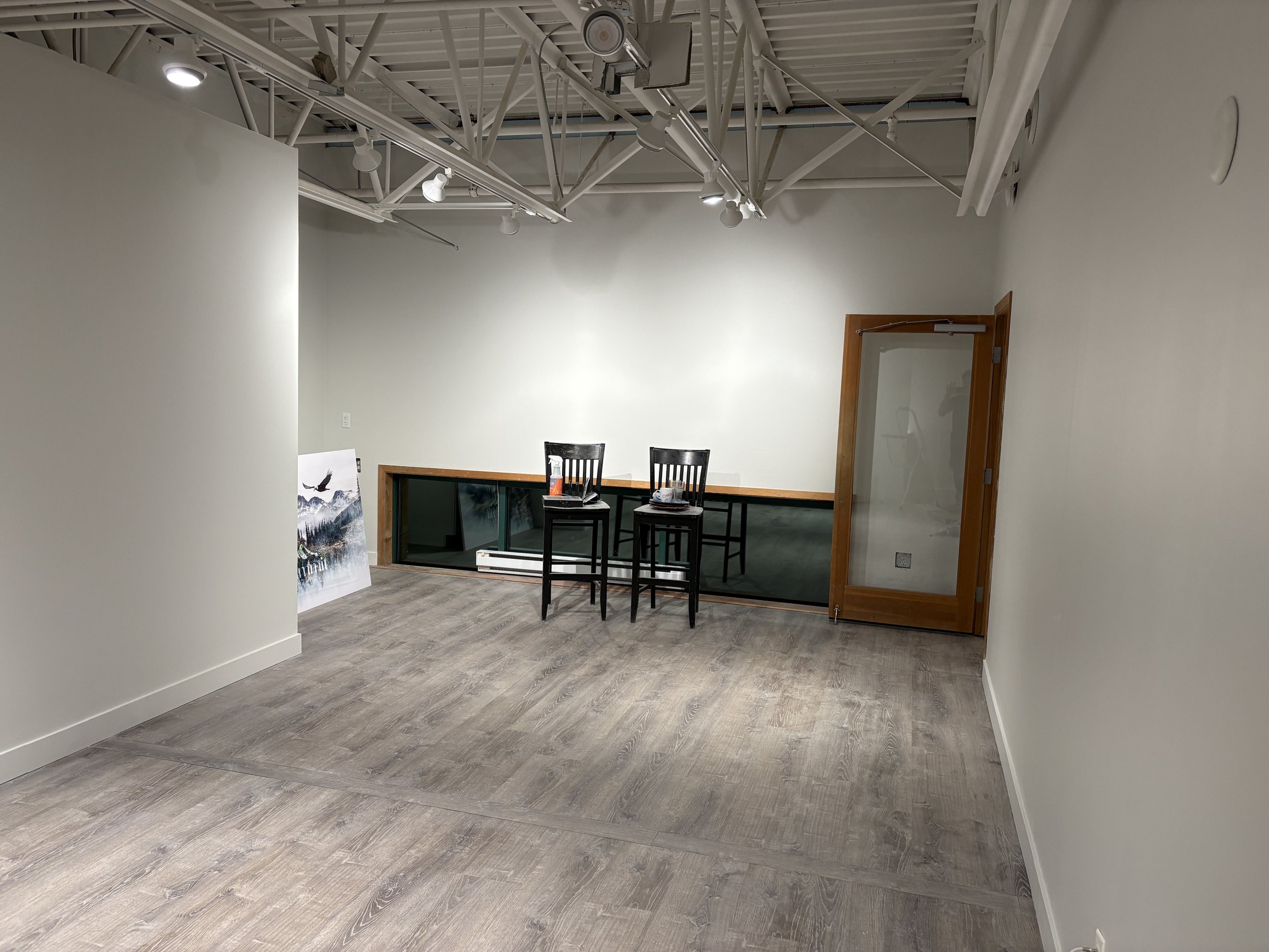 An empty room with white walls, gray wood flooring, and a high ceiling with visible white structural beams and lighting. Three black chairs are placed facing a windowed platform with a glass railing. A landscape painting of mountains and trees with a