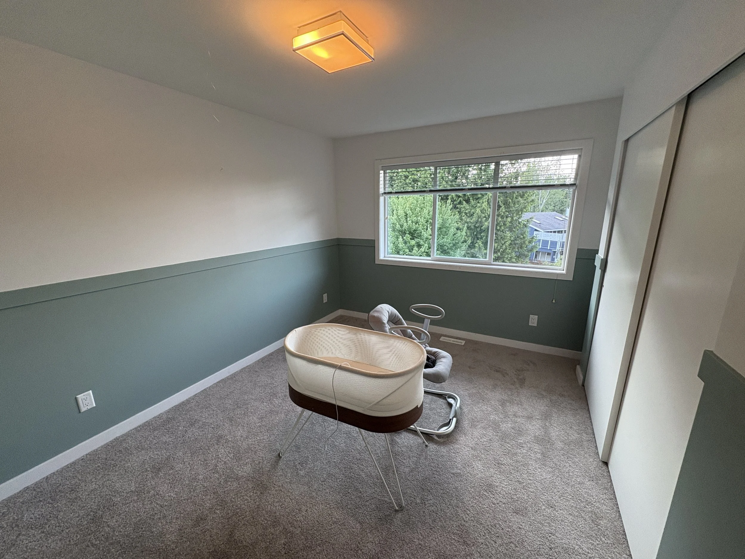 Empty room with a window, carpeted floor, and a white and brown bassinet with a grey recliner chair nearby.