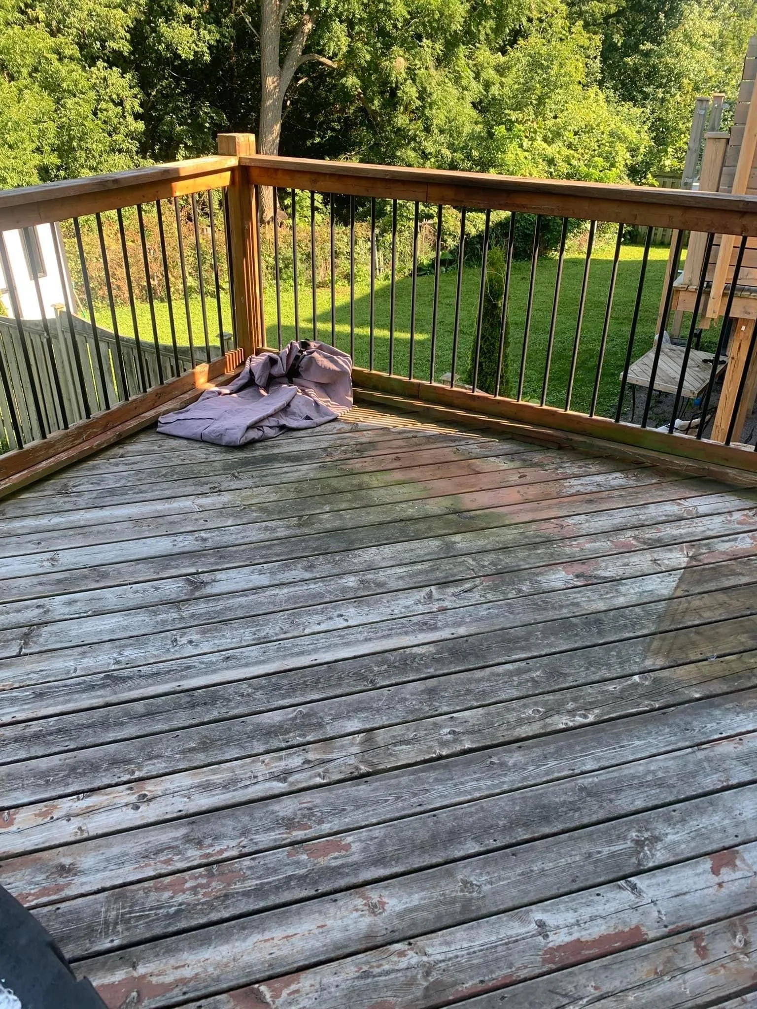 Outdoor wooden deck with peeling paint and railing, view of grassy yard and trees, folded purple tarp in corner.