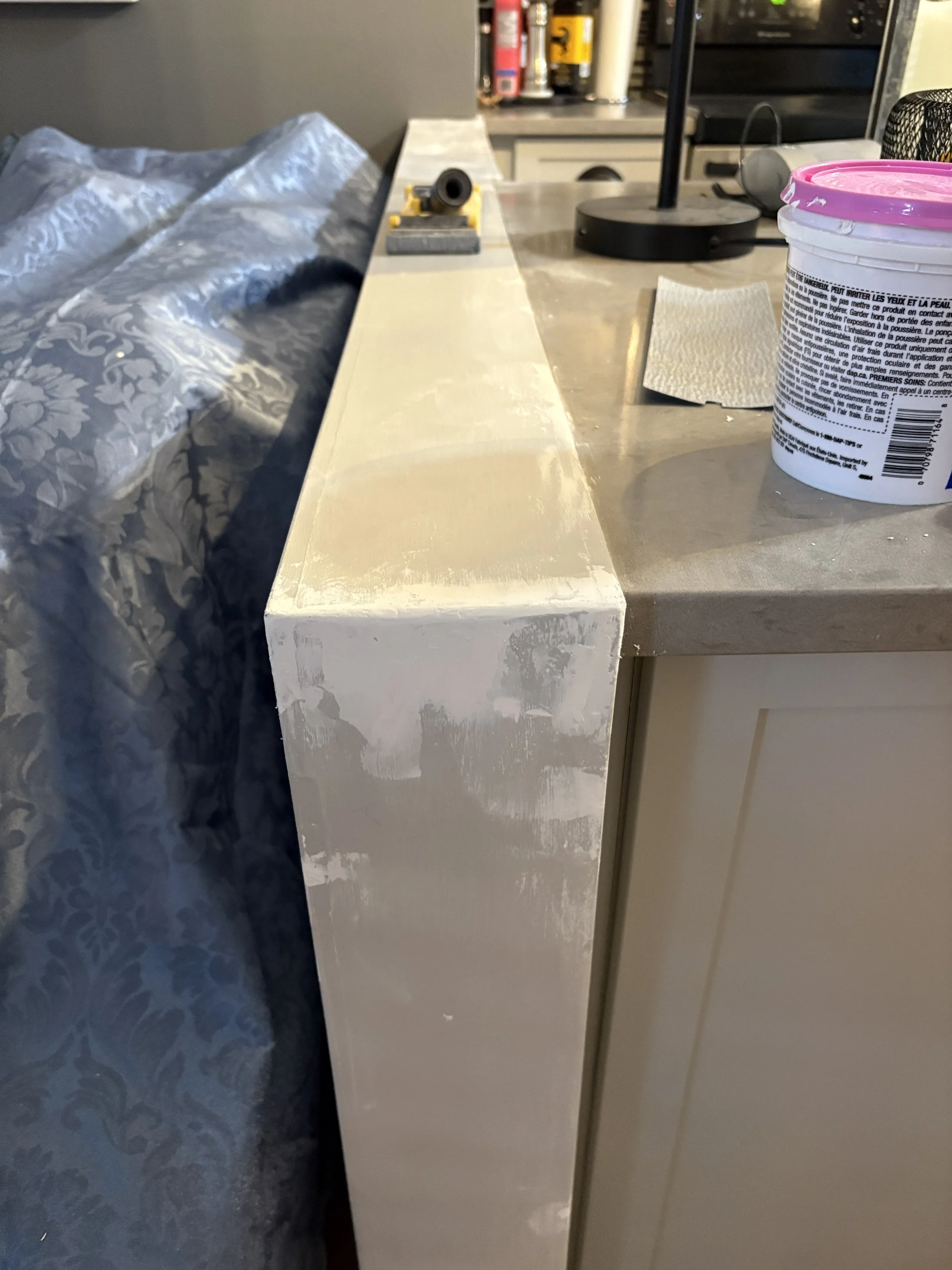 Home improvement project showing a partially sanded wooden surface with a sanding block and a container of spackling paste on a counter.
