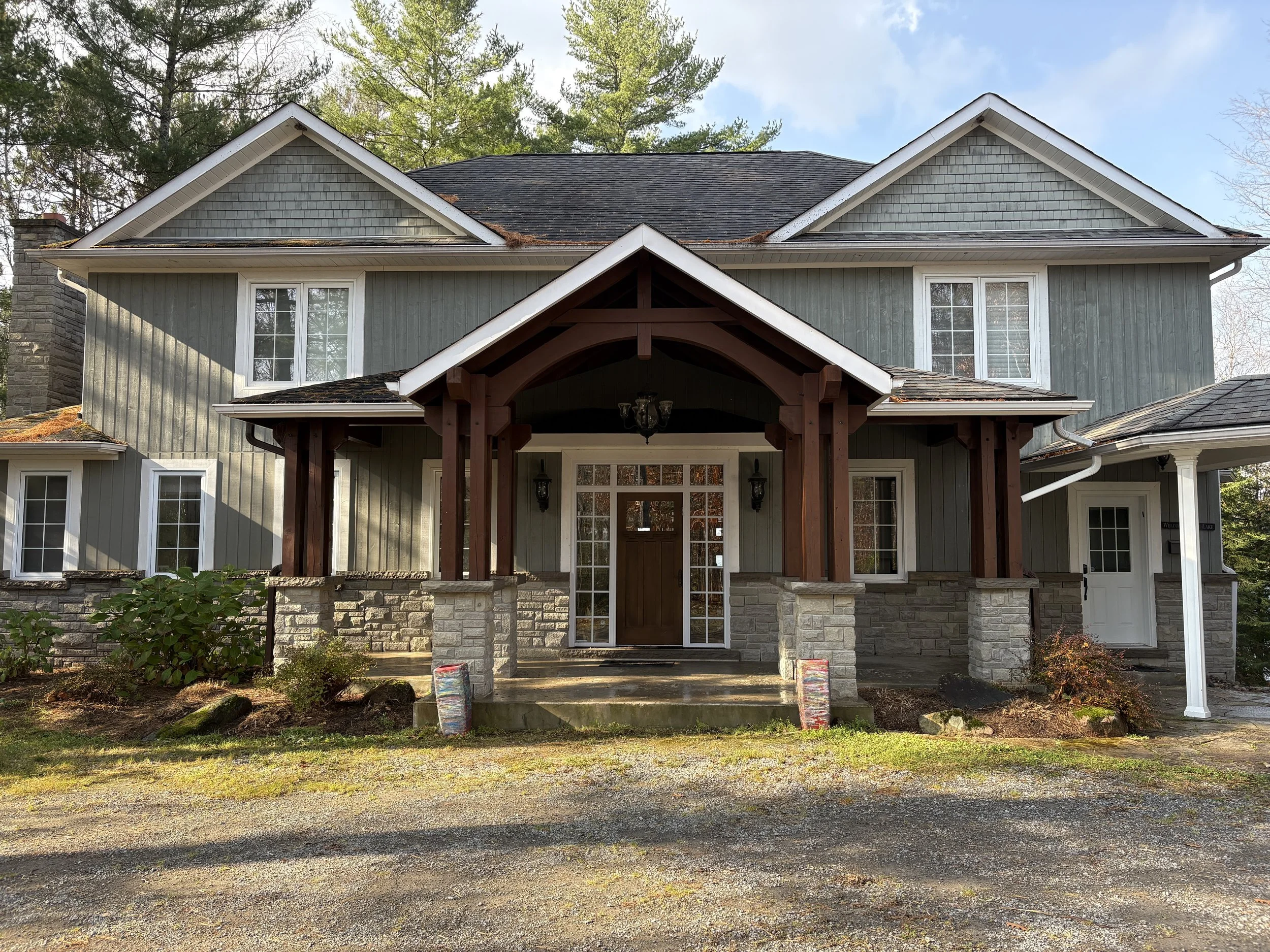 Front view of a two-story house with gray siding, stone accents, white trim, and a covered porch supported by wooden columns. The house has multiple windows, a dark shingled roof, and surrounding greenery.