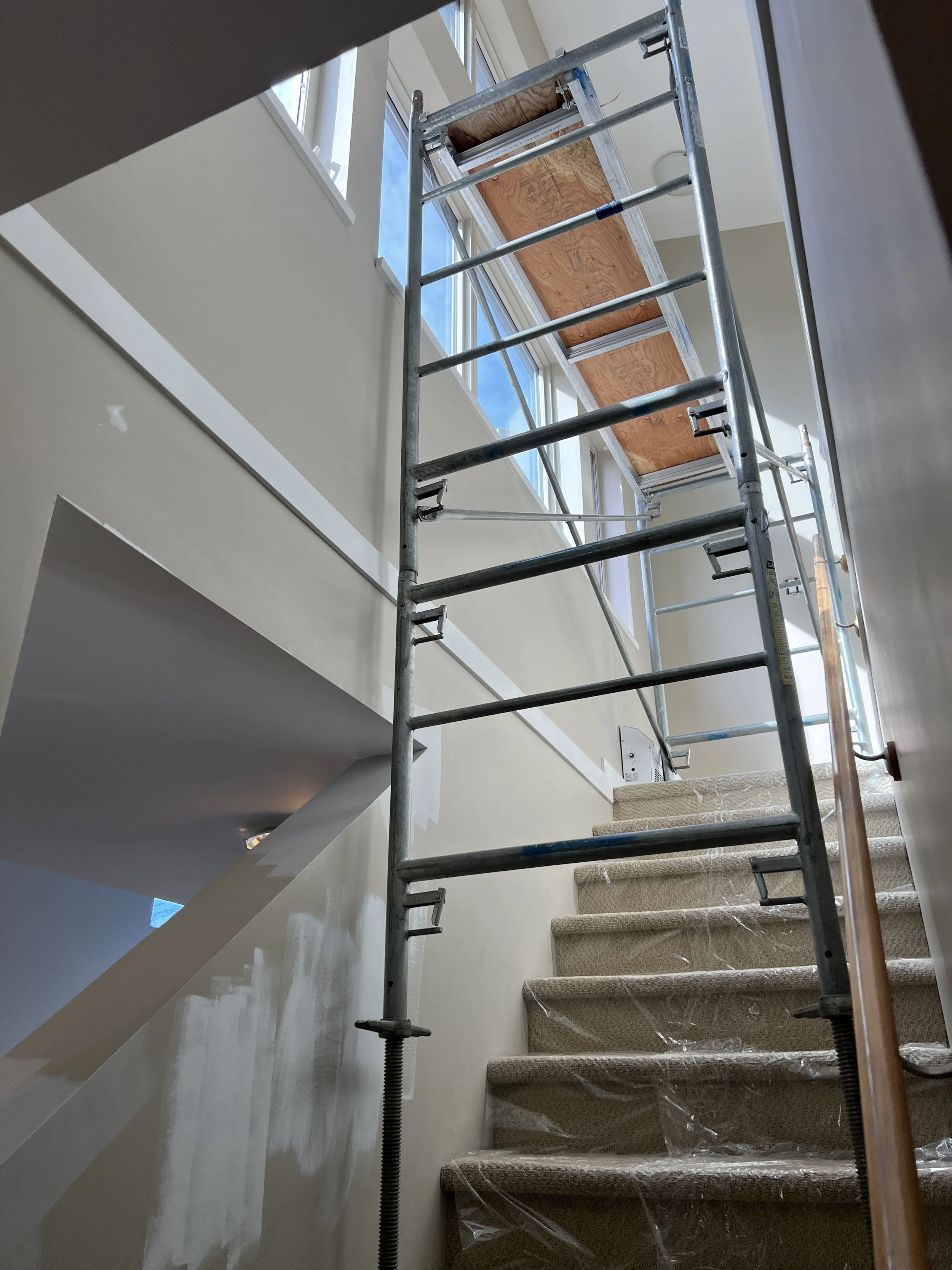 Indoor construction scene with scaffolding setup on a staircase for reaching high walls or ceilings, plastic covering on carpeted steps, and partially painted walls, next to windows with visible blue sky.